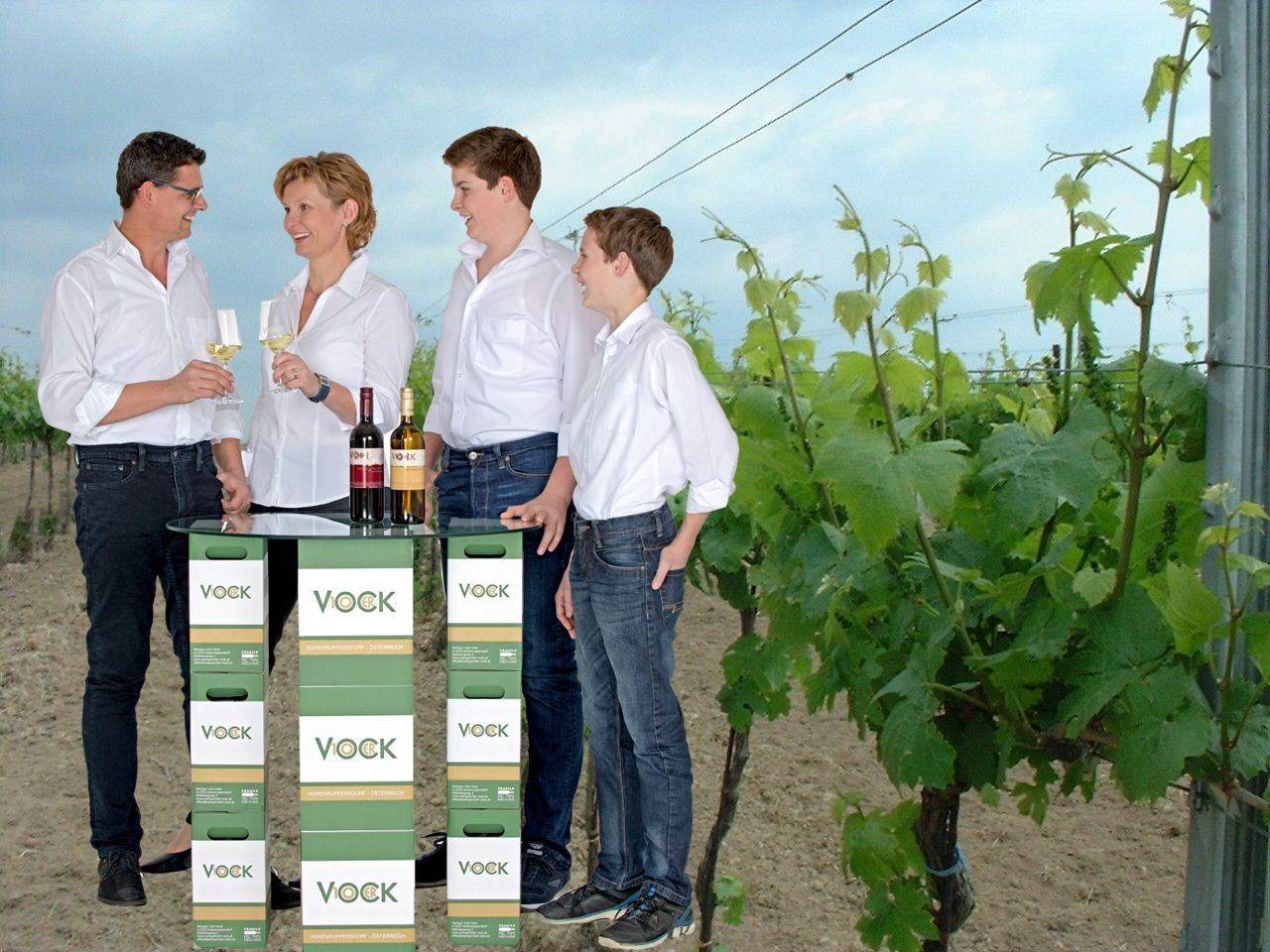 Vier Personen stehen in einem Weinberg und stoßen mit Weingläsern an. Vor ihnen ein Tisch aus Vock-Weinkartons.