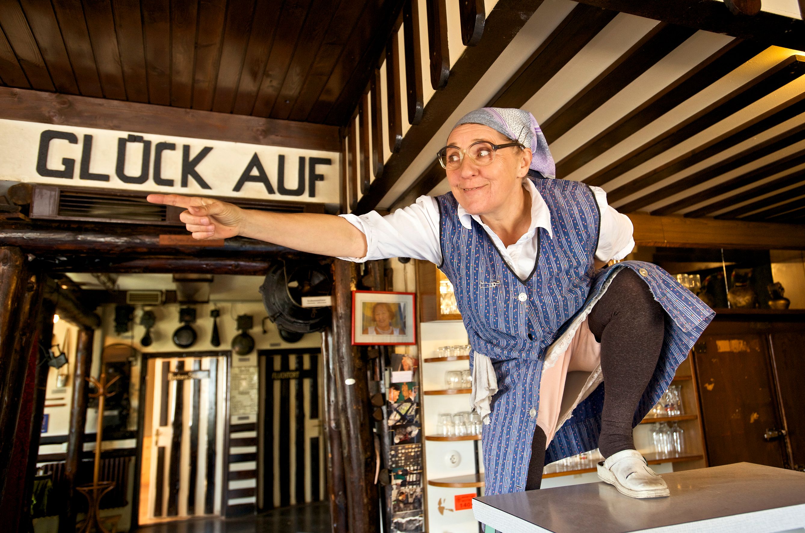 Eine Frau in traditioneller Kleidung zeigt in einem rustikalen Raum auf etwas. Über ihr steht "Glück Auf".