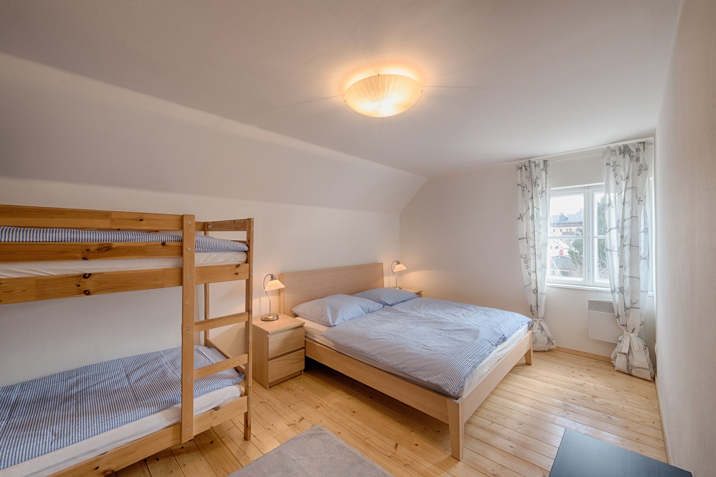Helles Schlafzimmer mit Doppelbett und Etagenbett, Holzboden und Fenster mit Vorhängen.