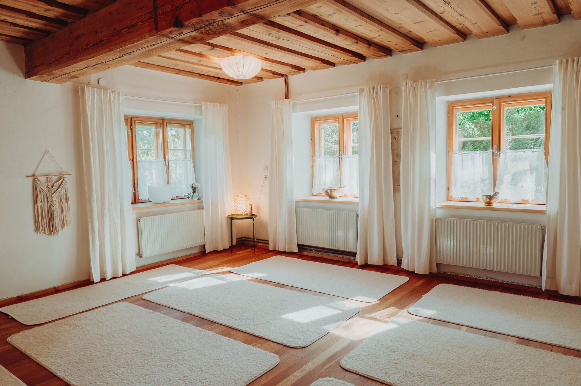 Ein heller Yogaraum mit Holzboden, weißen Matten und großen Fenstern mit Vorhängen.