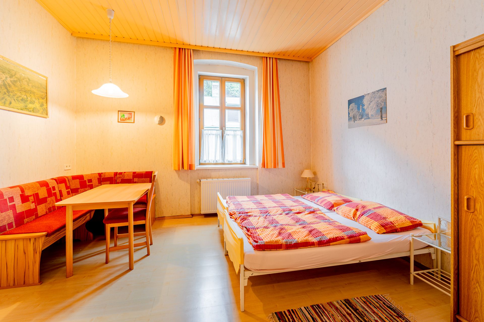 Gemütliches Schlafzimmer mit Doppelbett, Holzmöbeln und Esstisch. Helle Vorhänge und warme Farben dominieren den Raum.