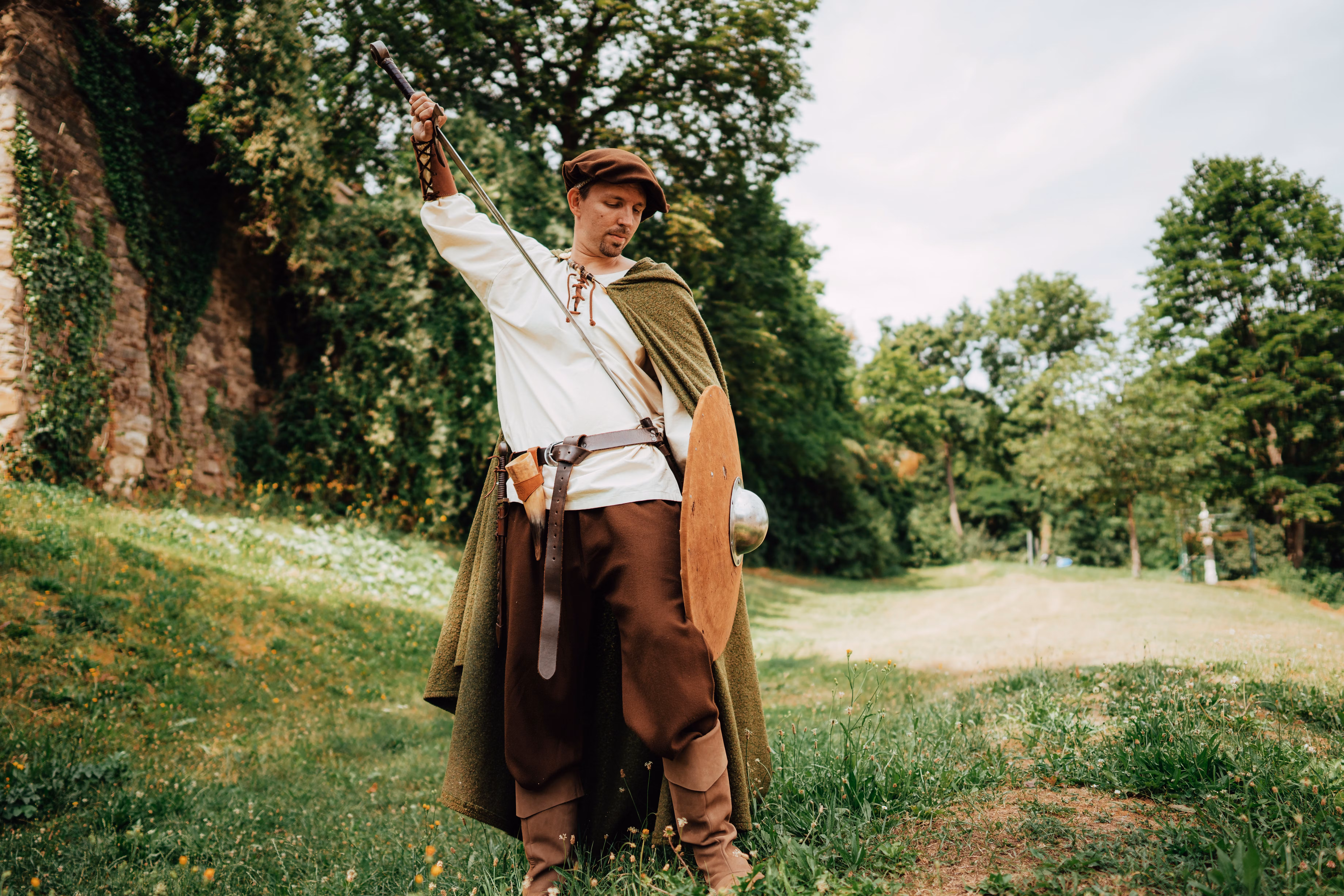 Ein Mann in mittelalterlicher Kleidung mit Schwert und Schild steht auf einer Wiese vor einer bewachsenen Steinmauer.