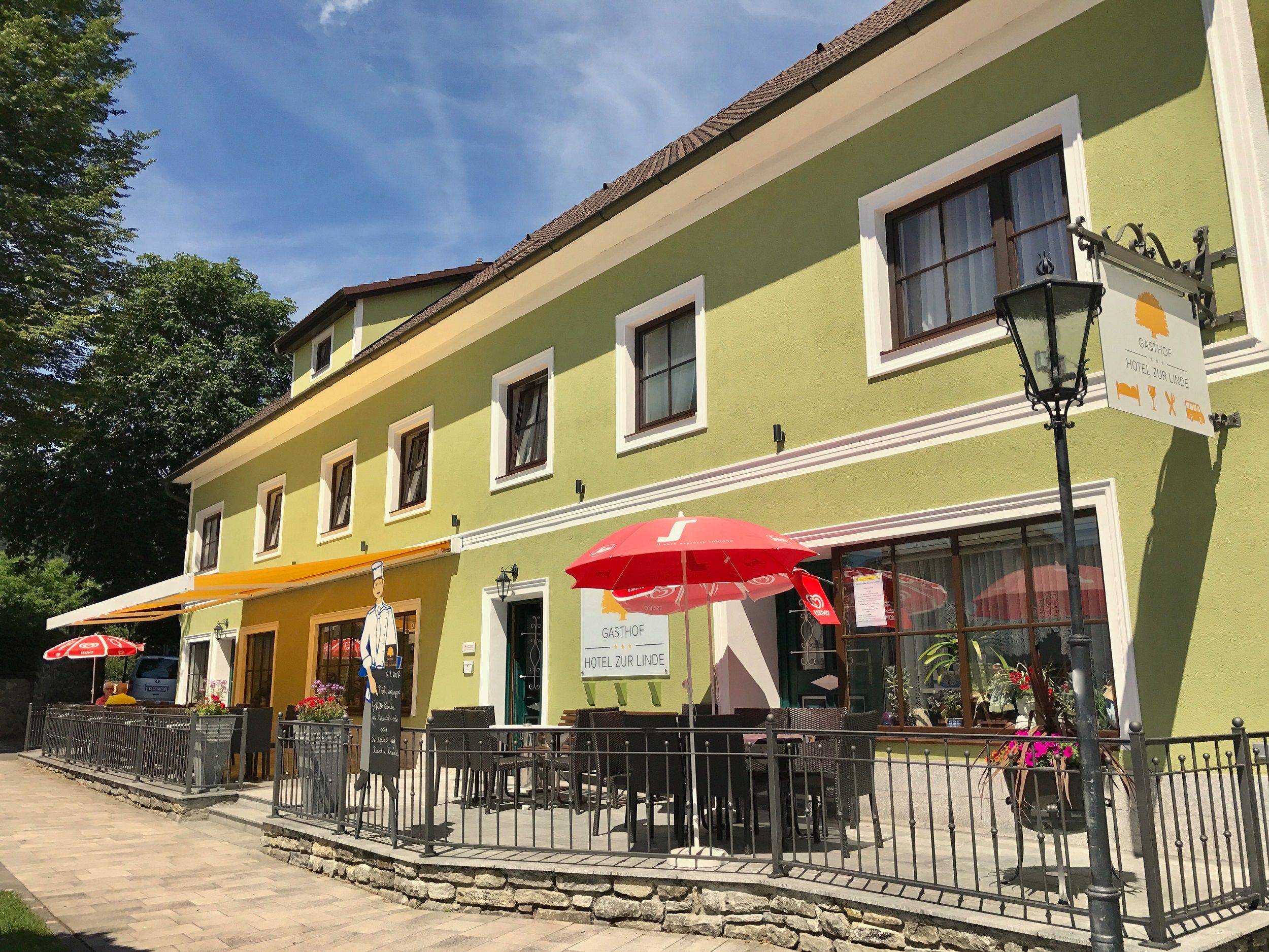Ein grüner Gasthof mit Terrasse und roten Sonnenschirmen, beschriftet mit 'Gasthof zur Linde'.