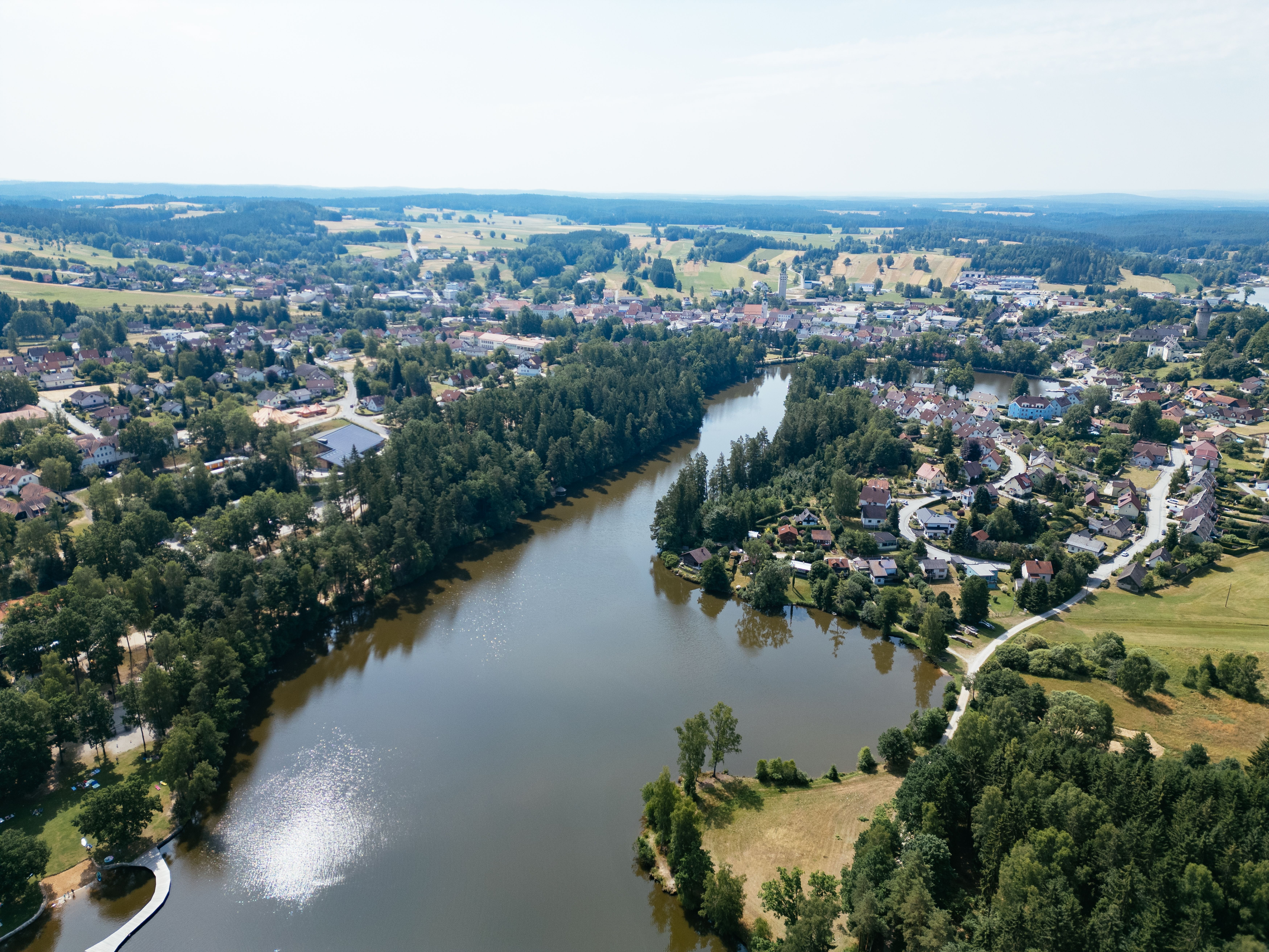 Luftaufnahme eines Baches, der durch eine von Wald und einer kleinen Siedlungen umgebene Landschaft fließt