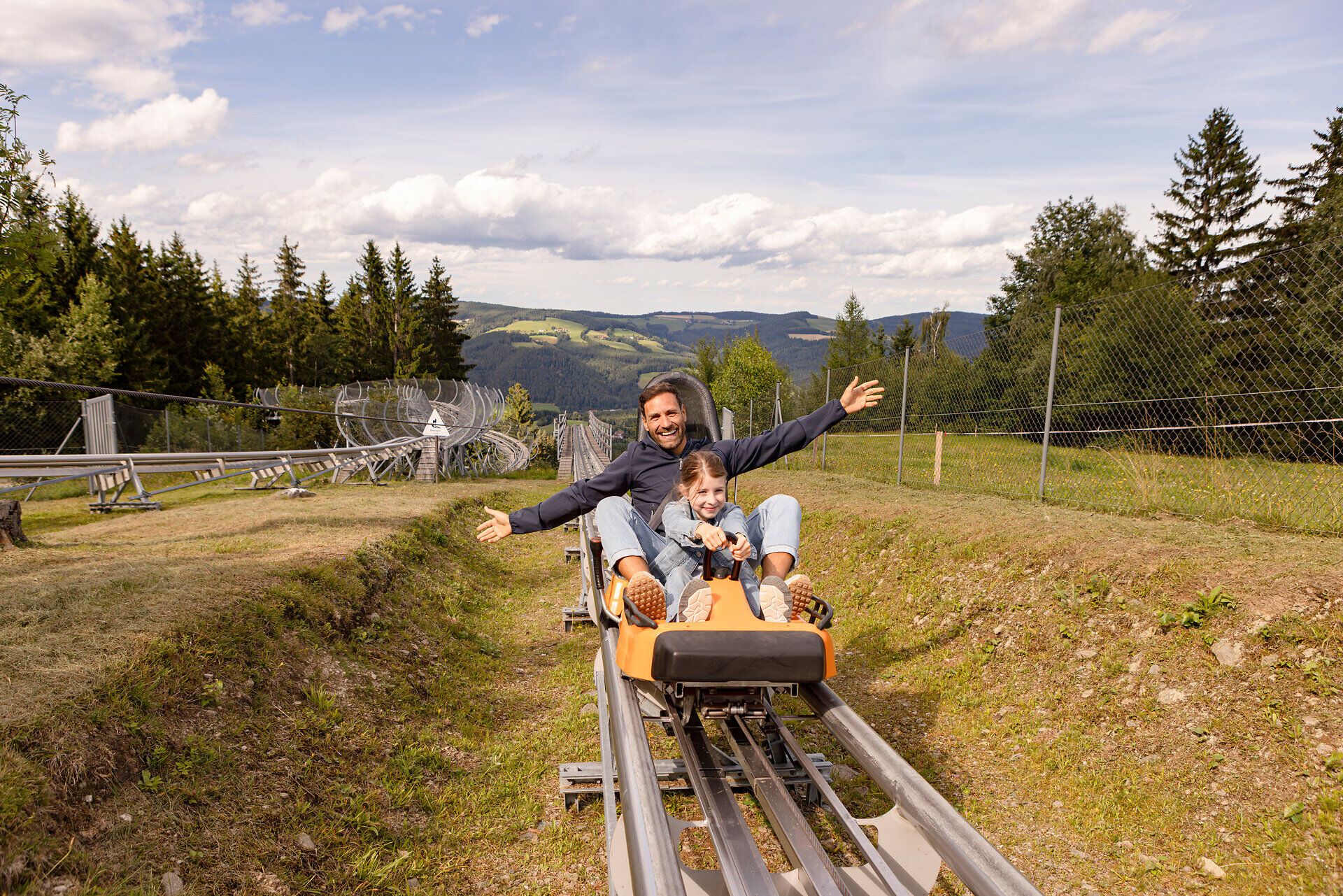 Ein fröhlicher Tag in der Natur, wo ein Vater mit seinem Kind auf einer Sommerrodelbahn die aufregende Abfahrt genießt. Umgeben von grünen Wäldern und sanften Hügeln, strahlt die Szene pure Lebensfreude und Abenteuerlust aus.
