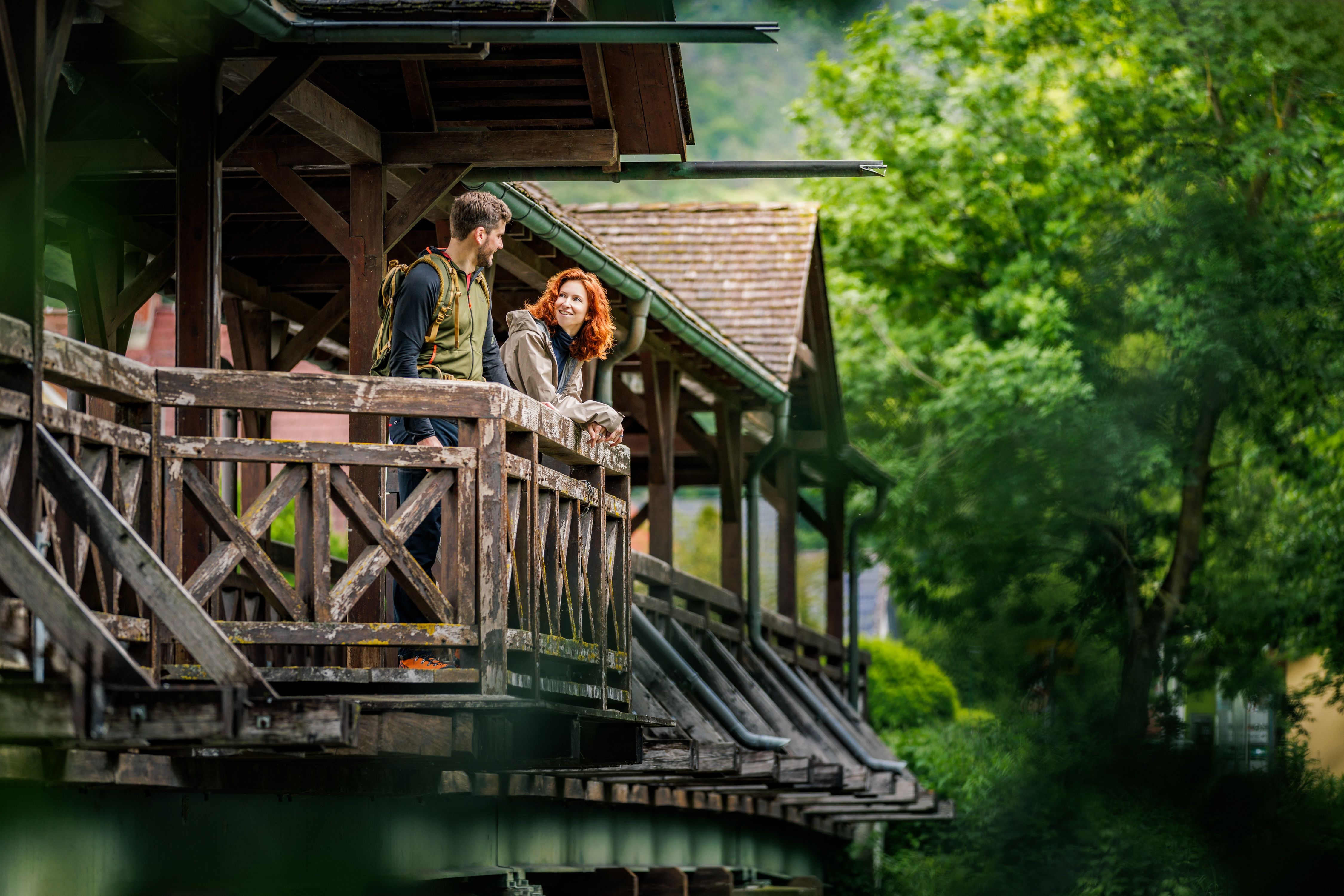 Ein Paar steht auf einer überdachten Holzbrücke und schaut sich an, umgeben von grüner Natur.