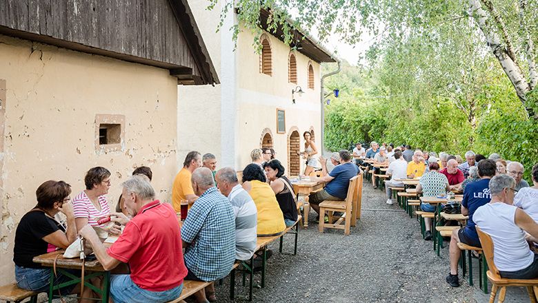 Menschen sitzen an langen Tischen im Freien bei einem Heurigen.