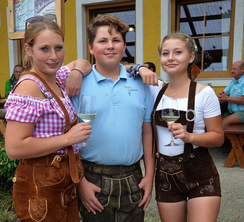 Drei junge Menschen in traditioneller bayerischer Kleidung mit Weingläsern vor einem gelben Gebäude.