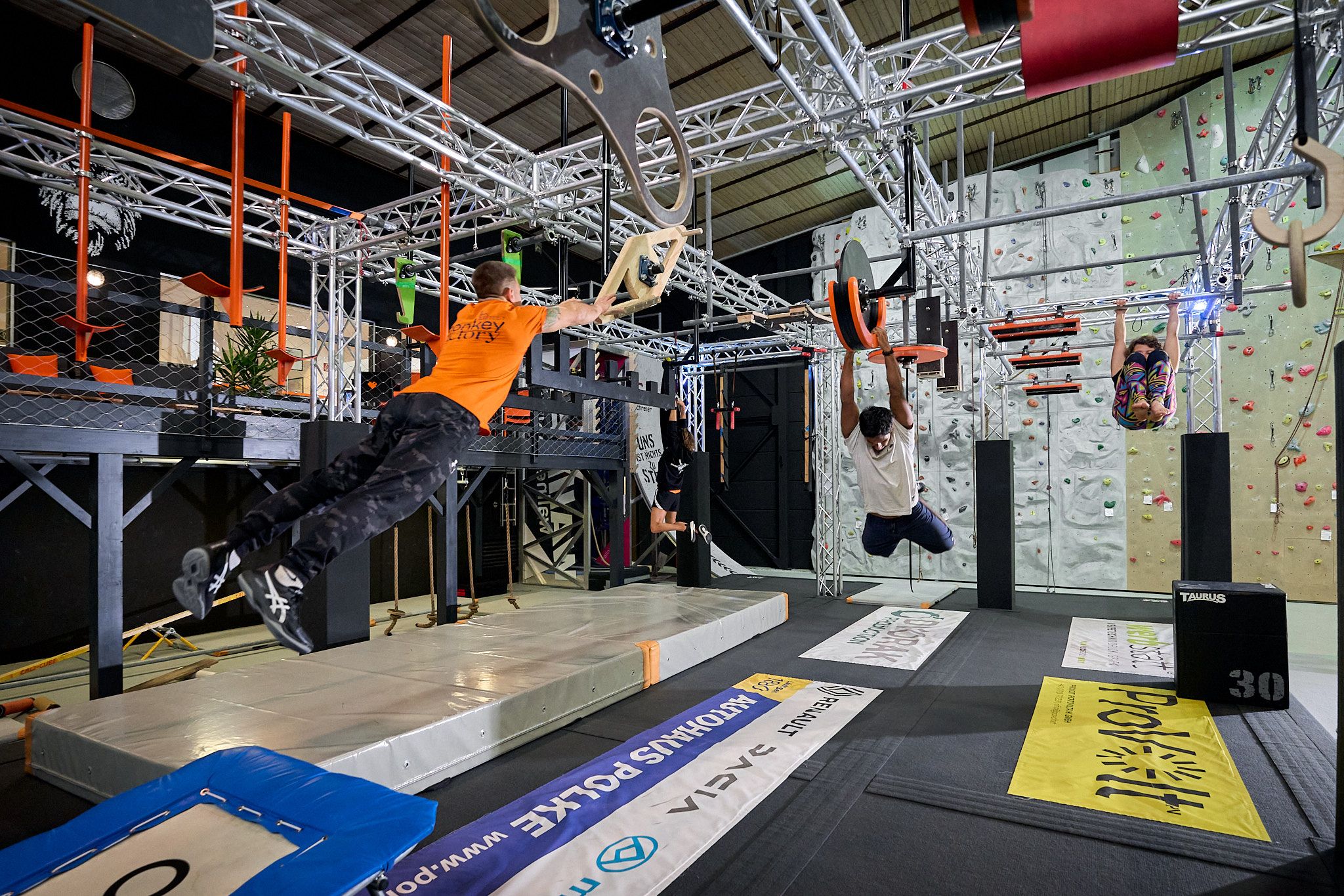 Menschen trainieren in einem Indoor-Kletterpark mit verschiedenen Hindernissen und Kletterwänden.