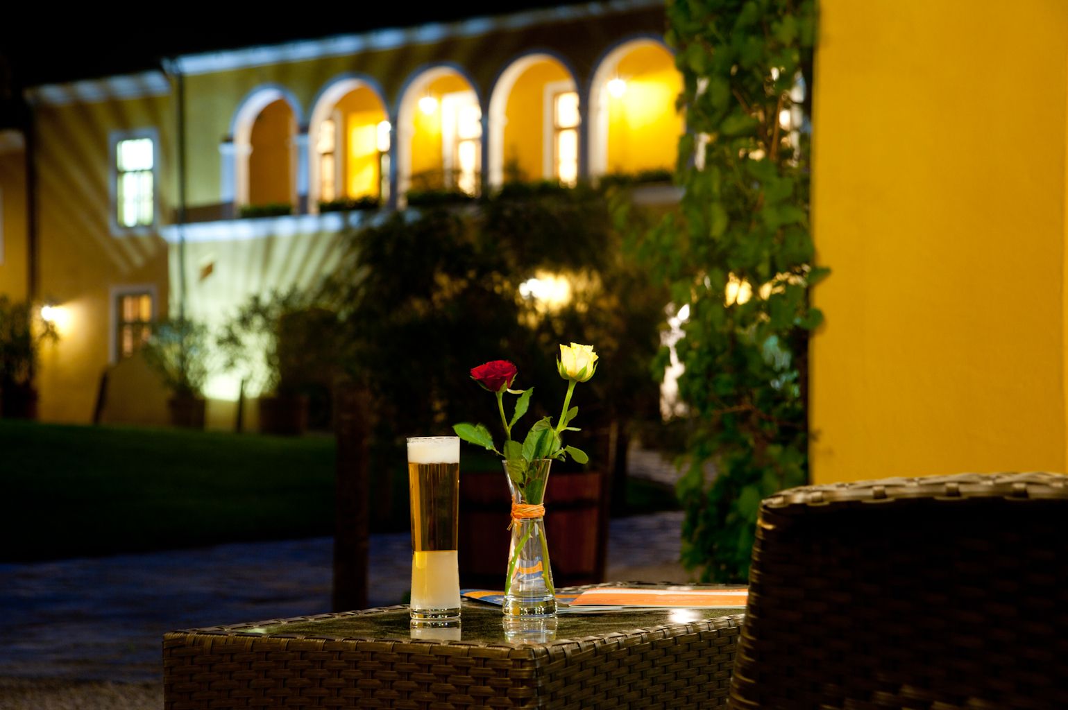 Ein Tisch mit einem Glas Bier und zwei Rosen in einer Vase, im Hintergrund ein beleuchtetes Gebäude mit Bögen.