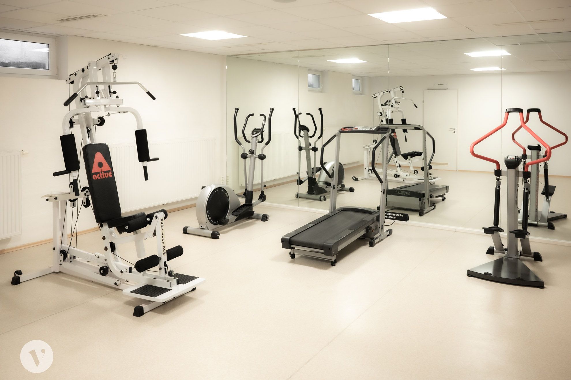 Ein Fitnessraum mit verschiedenen Trainingsgeräten wie Laufband, Heimtrainer und Kraftstation vor einem großen Spiegel.