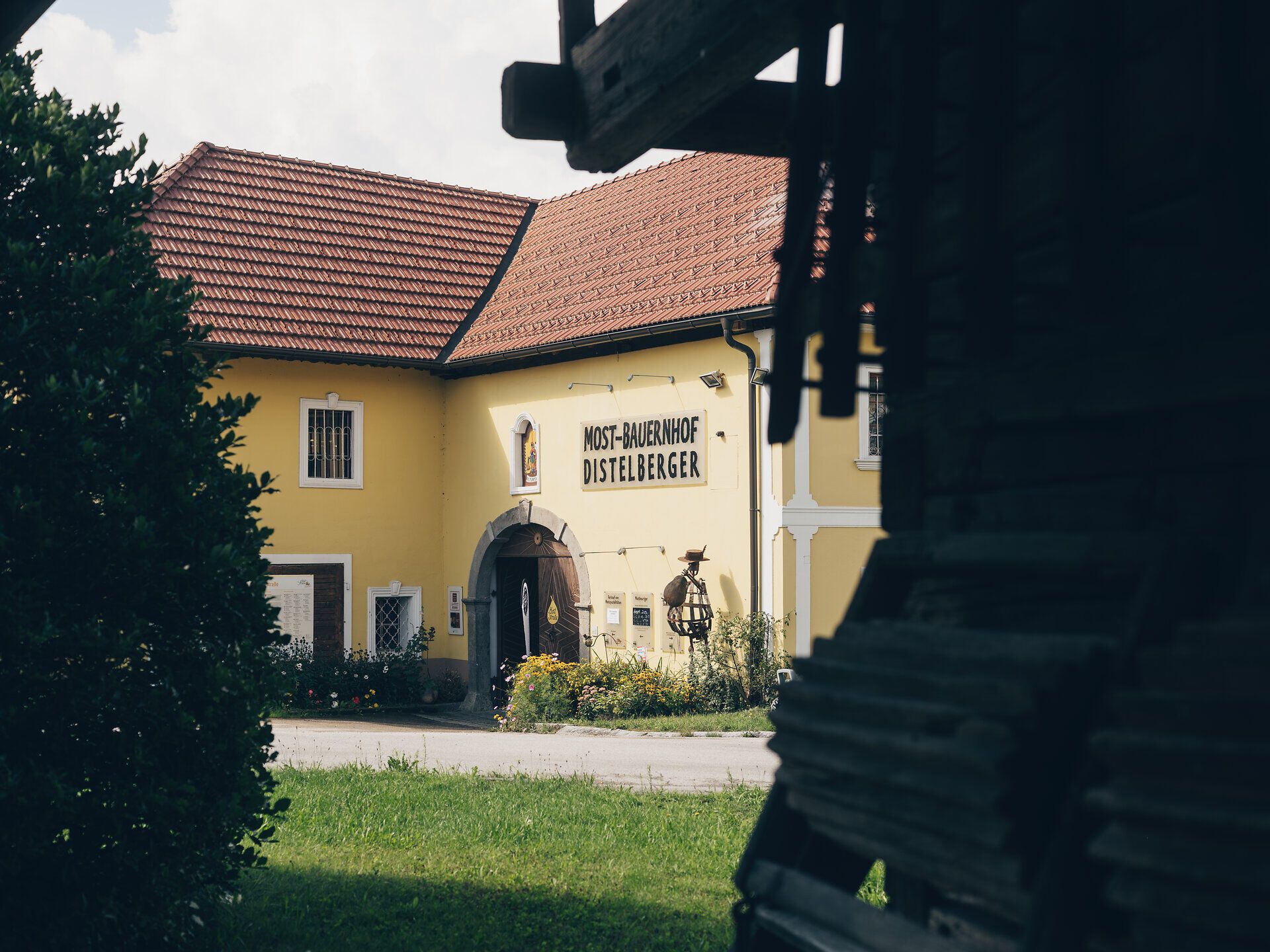 Ein malerischer GenussBauernhof mit strahlend gelben Wänden und einem einladenden Eingang, umgeben von blühenden Gärten und saftigem Grün. Hier erleben Besucher die herzliche Gastfreundschaft und die köstlichen Aromen des Mostviertels, während sie die idyllische Landschaft genießen.
