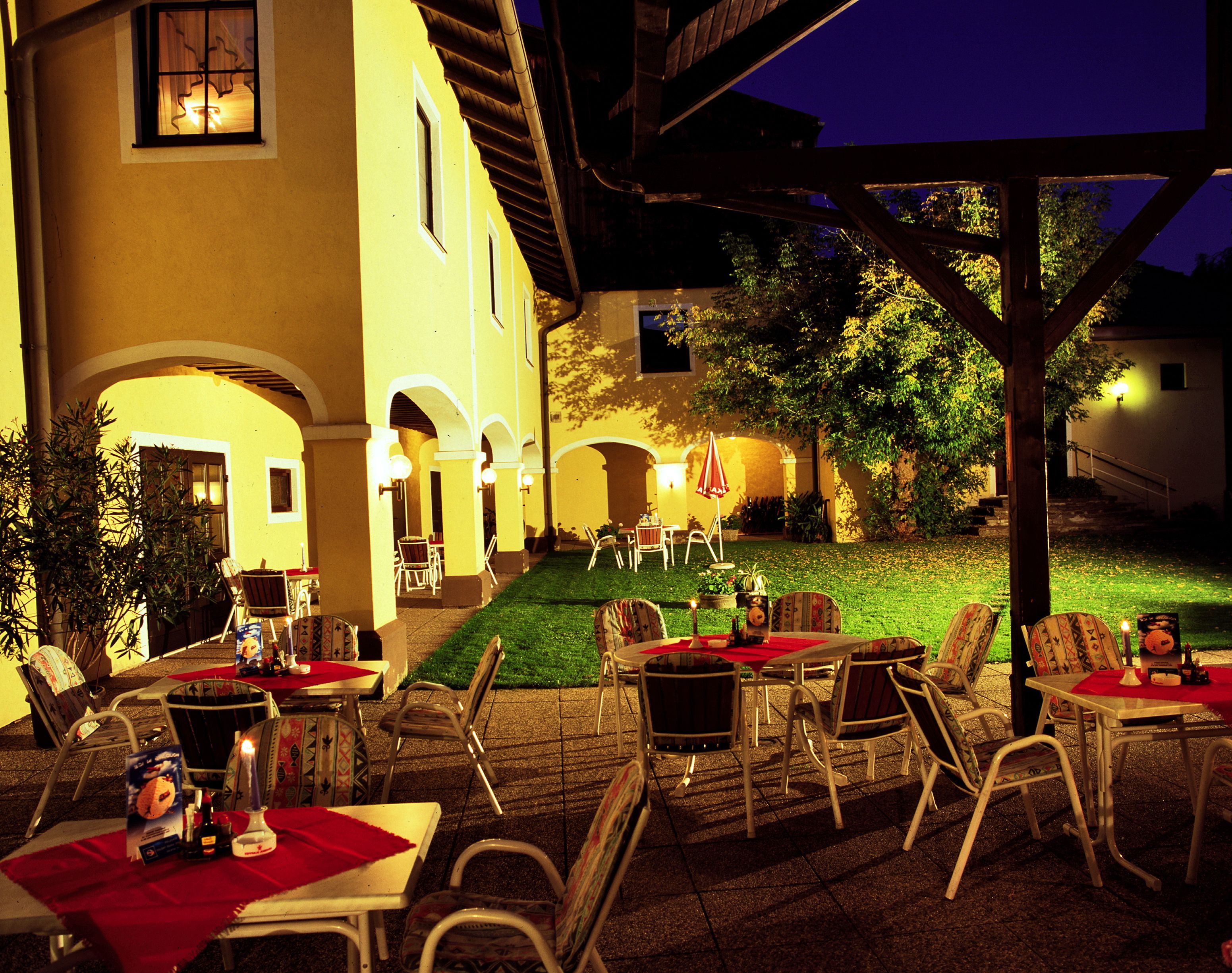 Beleuchteter Außenbereich eines Gasthofs mit gelben Wänden, Tischen und Stühlen auf einer Terrasse bei Nacht.