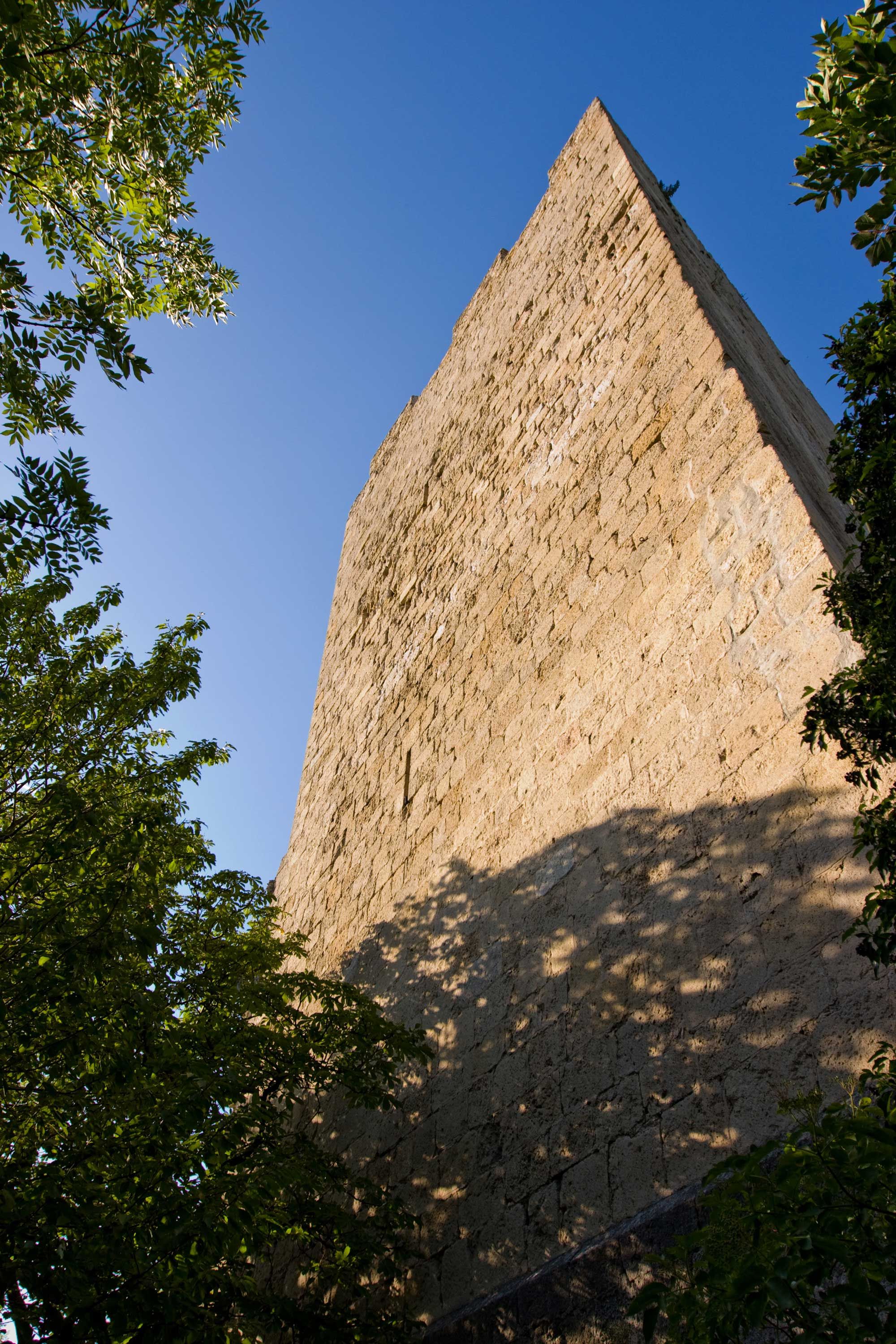 Nahaufnahme eines hohen, alten Burgturms aus Stein, umgeben von Bäumen, unter blauem Himmel.