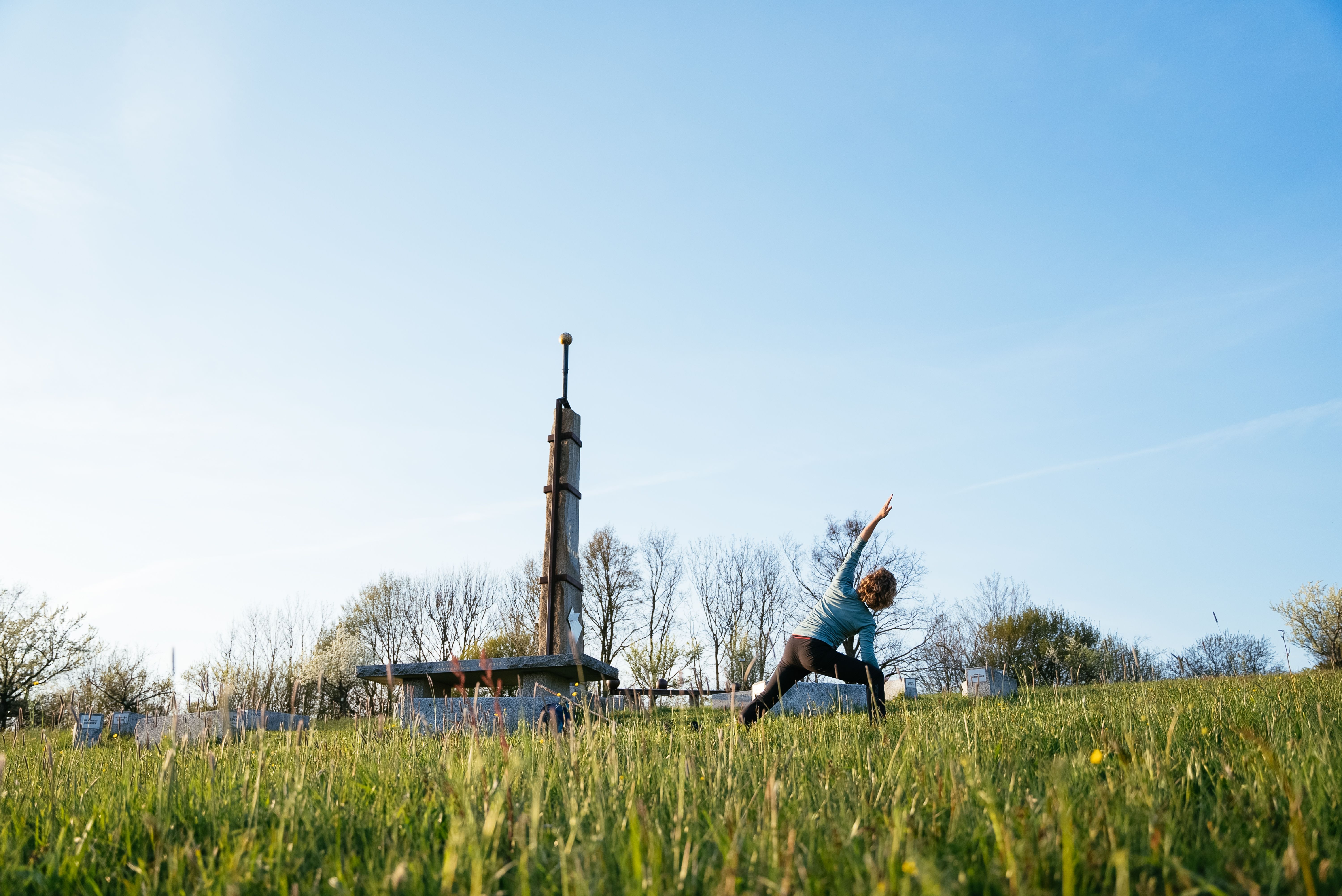 Frau macht auf einer sonnigen Wiese eine Yoga-Dehnung neben einem hohen Holzdenkmal, umgeben von Bäumen und weitem Himmel.