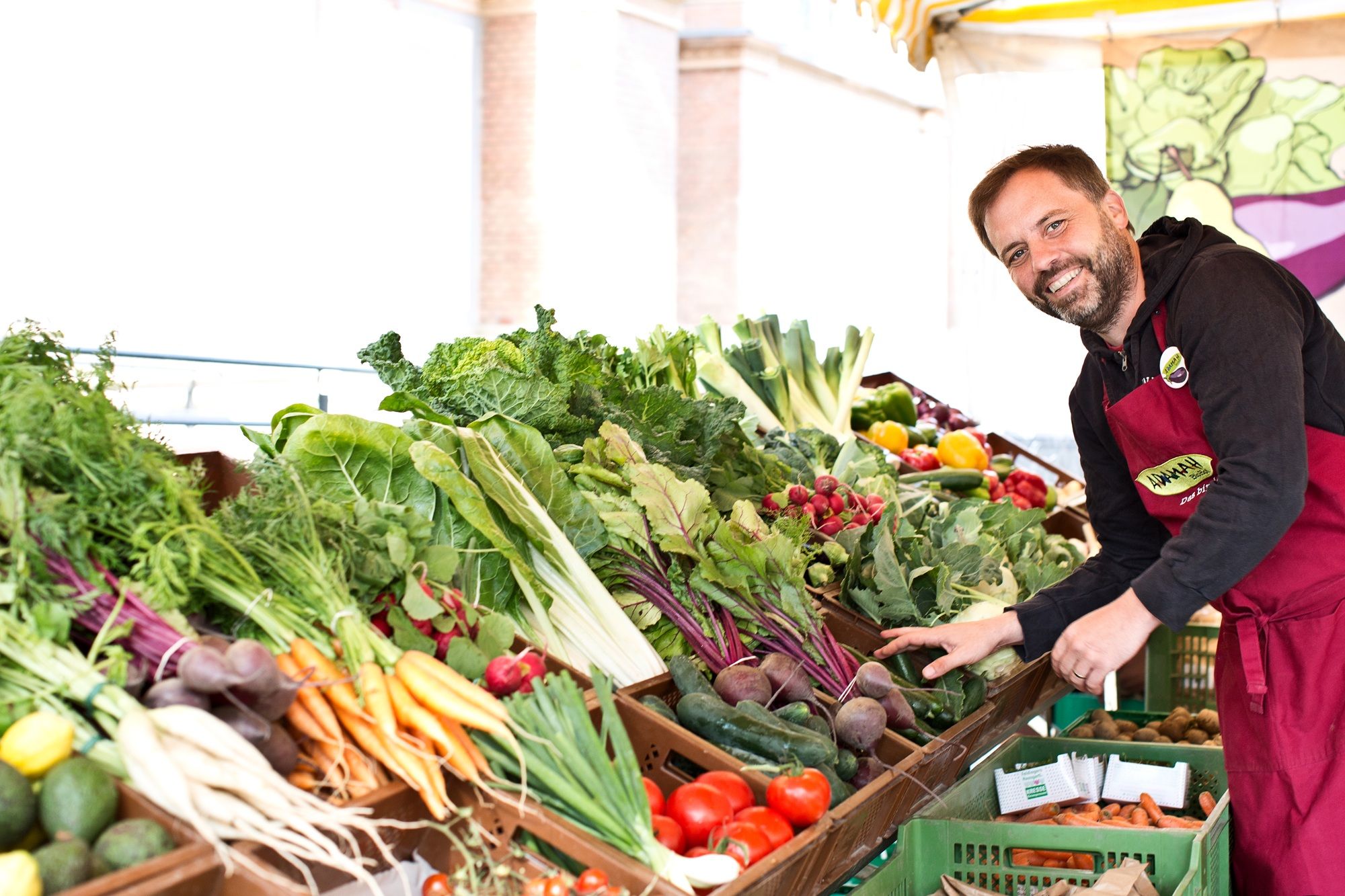 Ein lächelnder Marktverkäufer in roter Schürze steht hinter einem Stand mit frischem Gemüse.