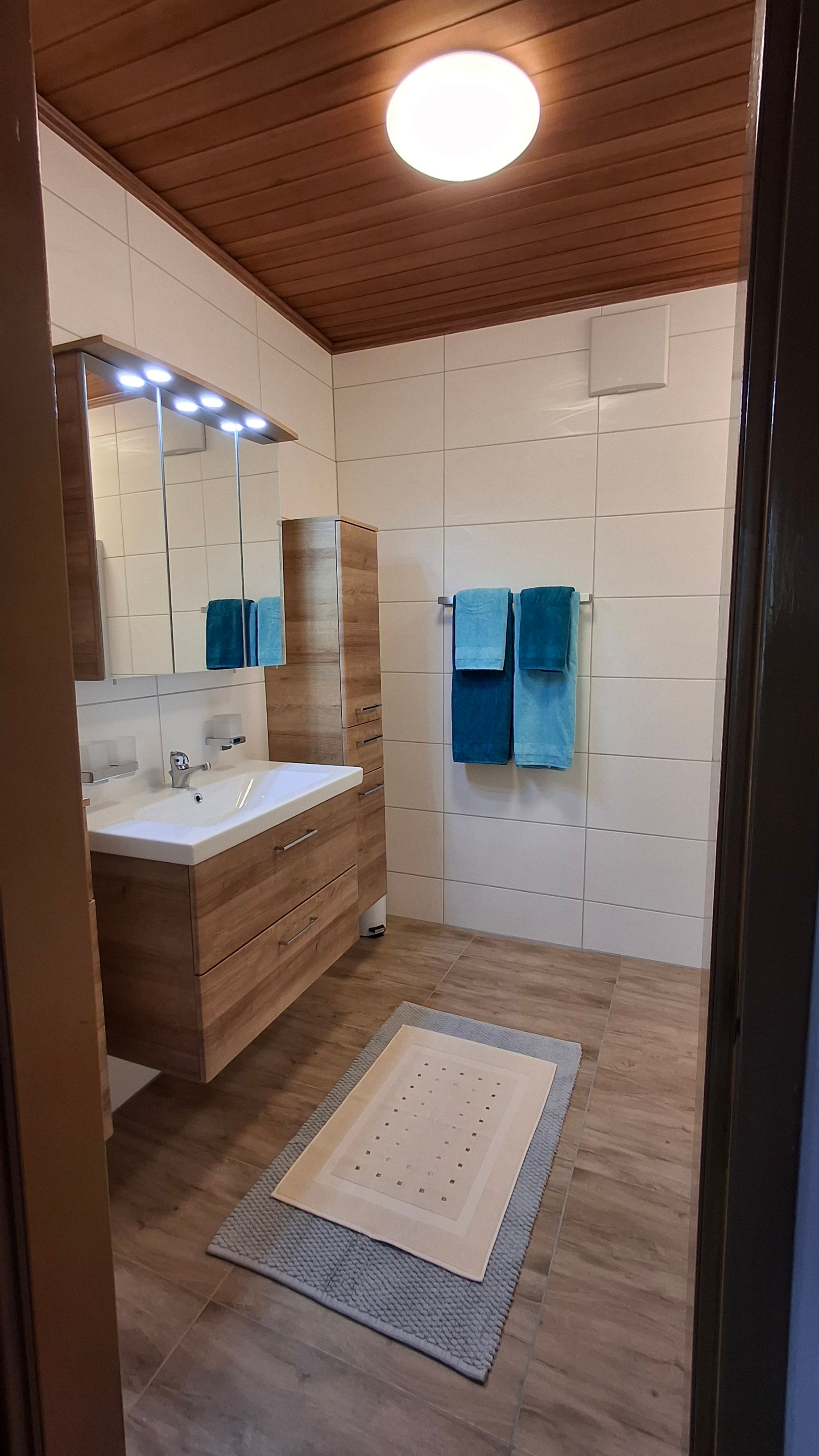 Modernes Badezimmer mit Holzdecke, Waschtisch, Spiegel, Handtüchern und Teppich.