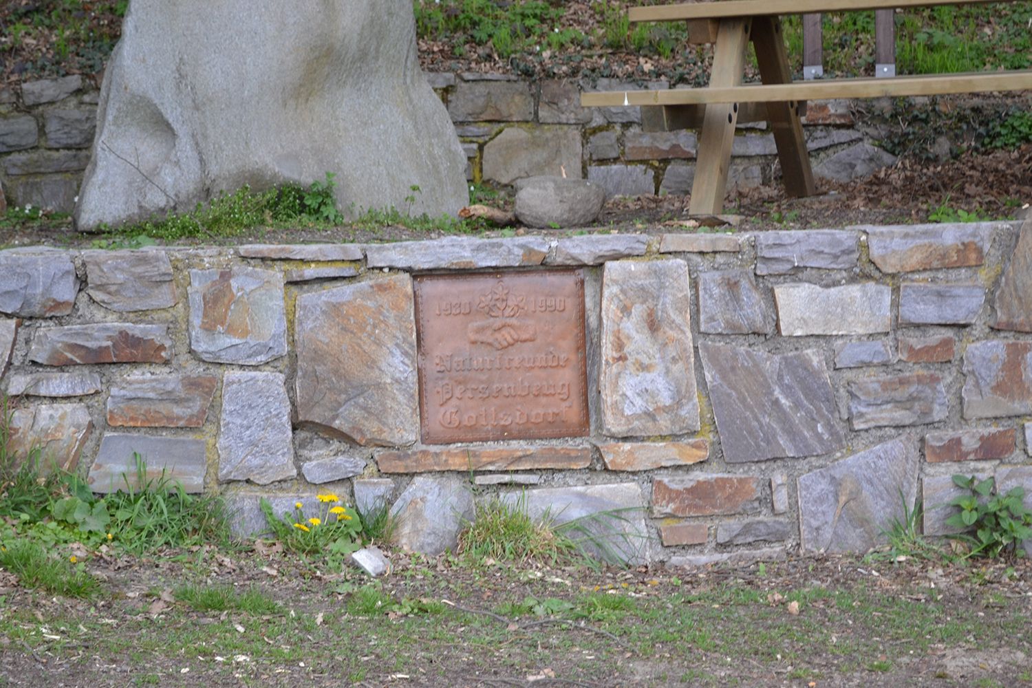 Steinmauer mit Gedenktafel und Picknicktisch im Hintergrund.