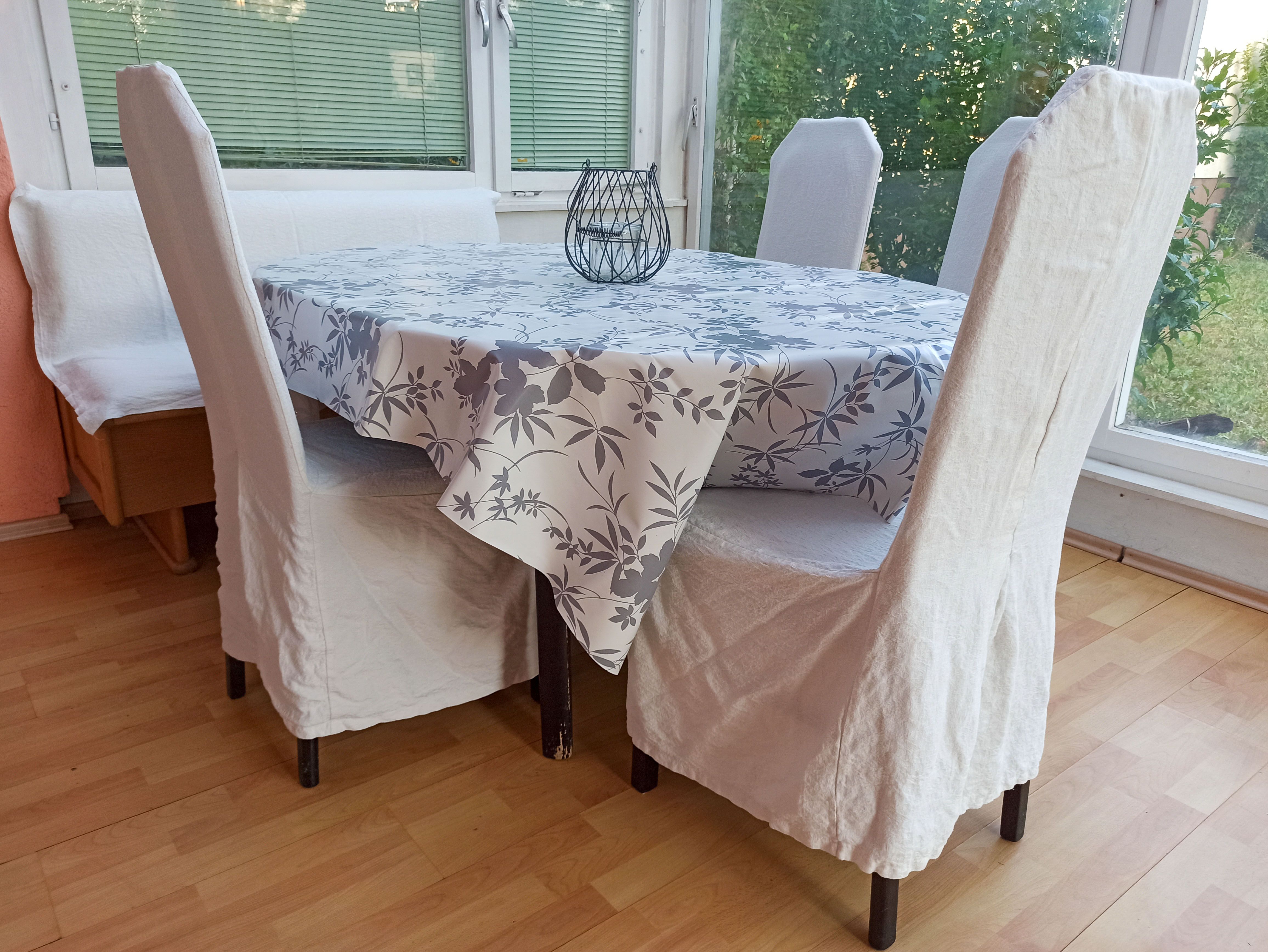 Essbereich mit Tisch, Stühlen und Bank, Tischdecke mit Blumenmuster, Fenster im Hintergrund.