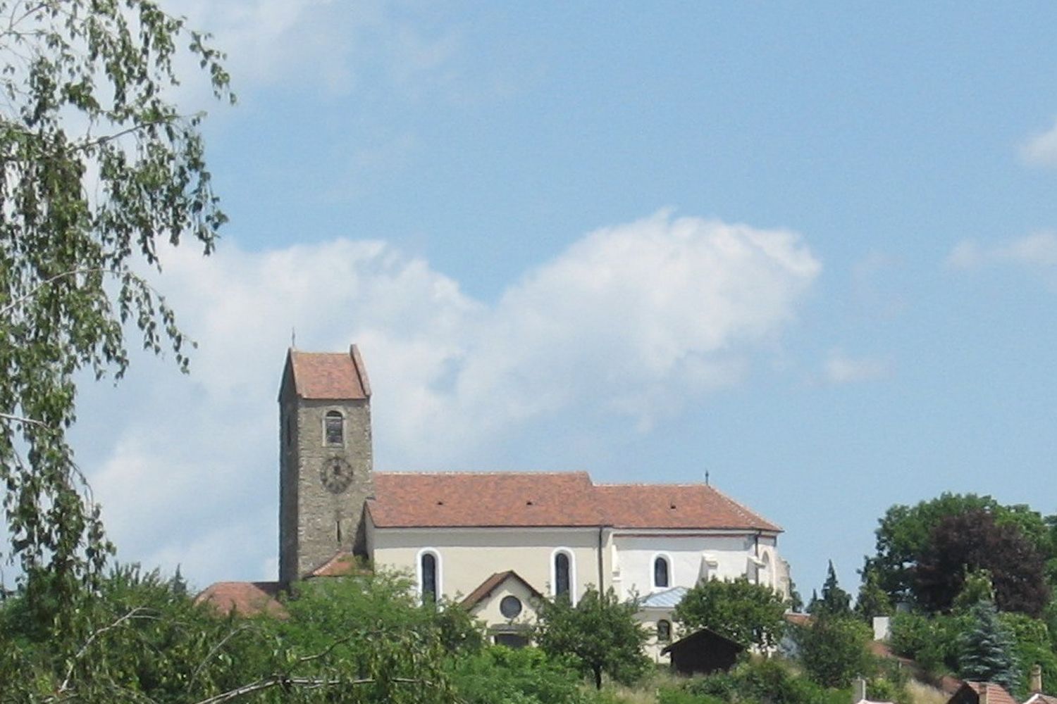 Kirche mit Turm und rotem Dach in Hohenwarth, umgeben von Bäumen und blauem Himmel.