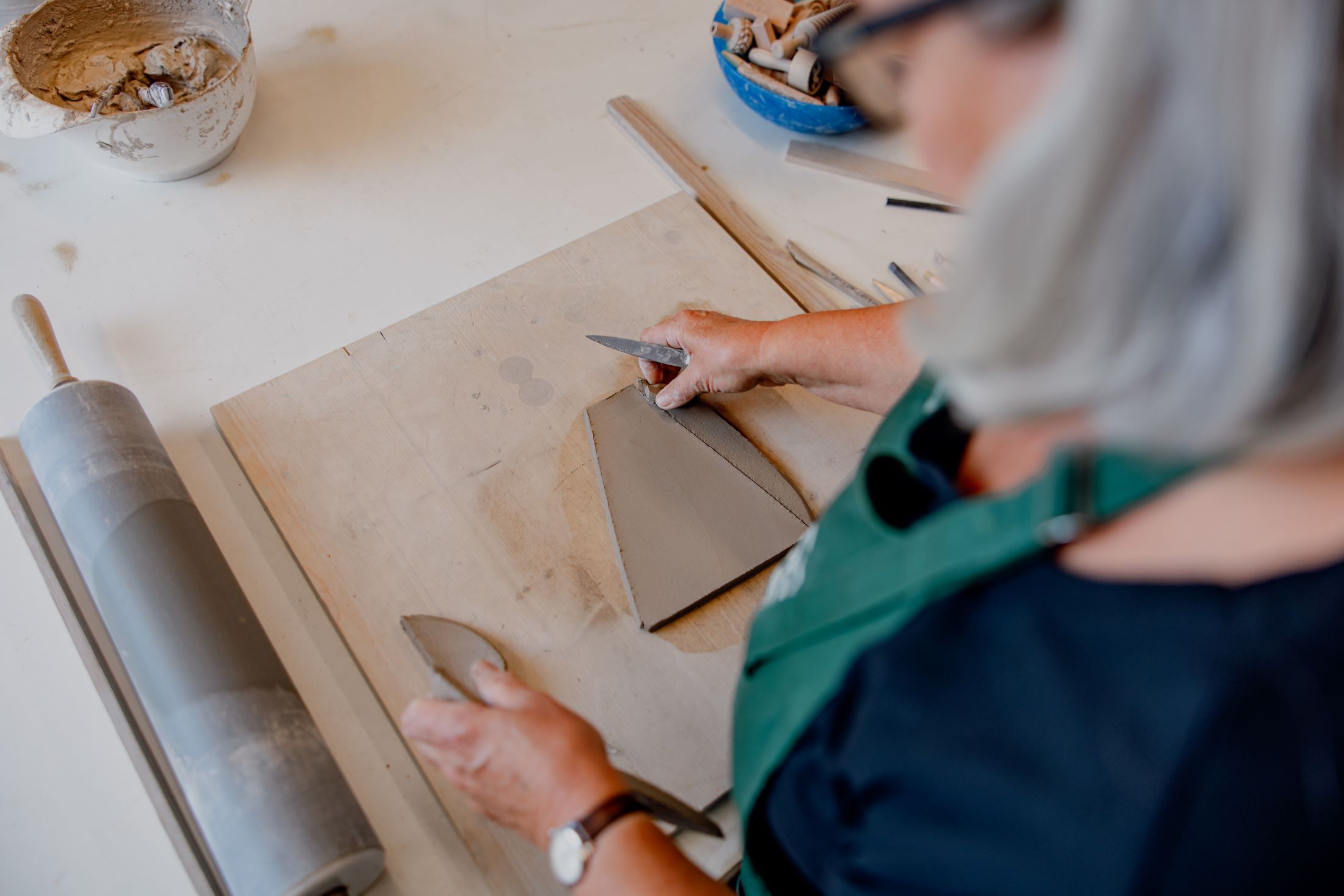 Person schneidet Ton auf einem Tisch mit einem Messer, umgeben von Töpferwerkzeugen.