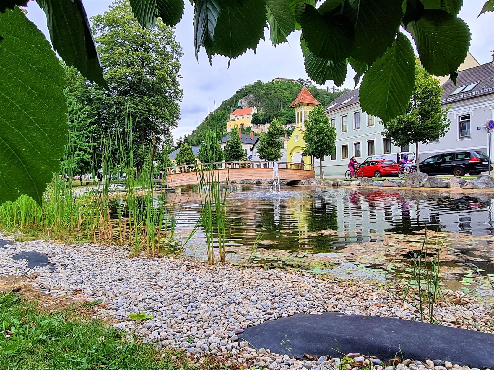Blick auf einen Teich mit Brücke und Bergkirche im Hintergrund, umgeben von Bäumen und Häusern.