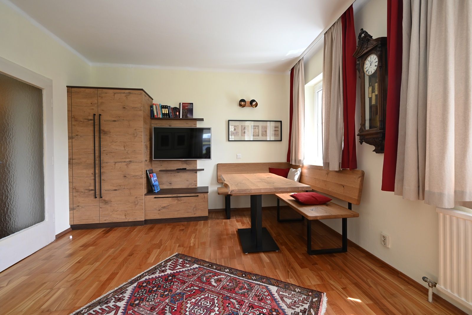 Gemütliches Wohnzimmer mit Holzmöbeln, einem Teppich, einer Eckbank und einer Wanduhr.