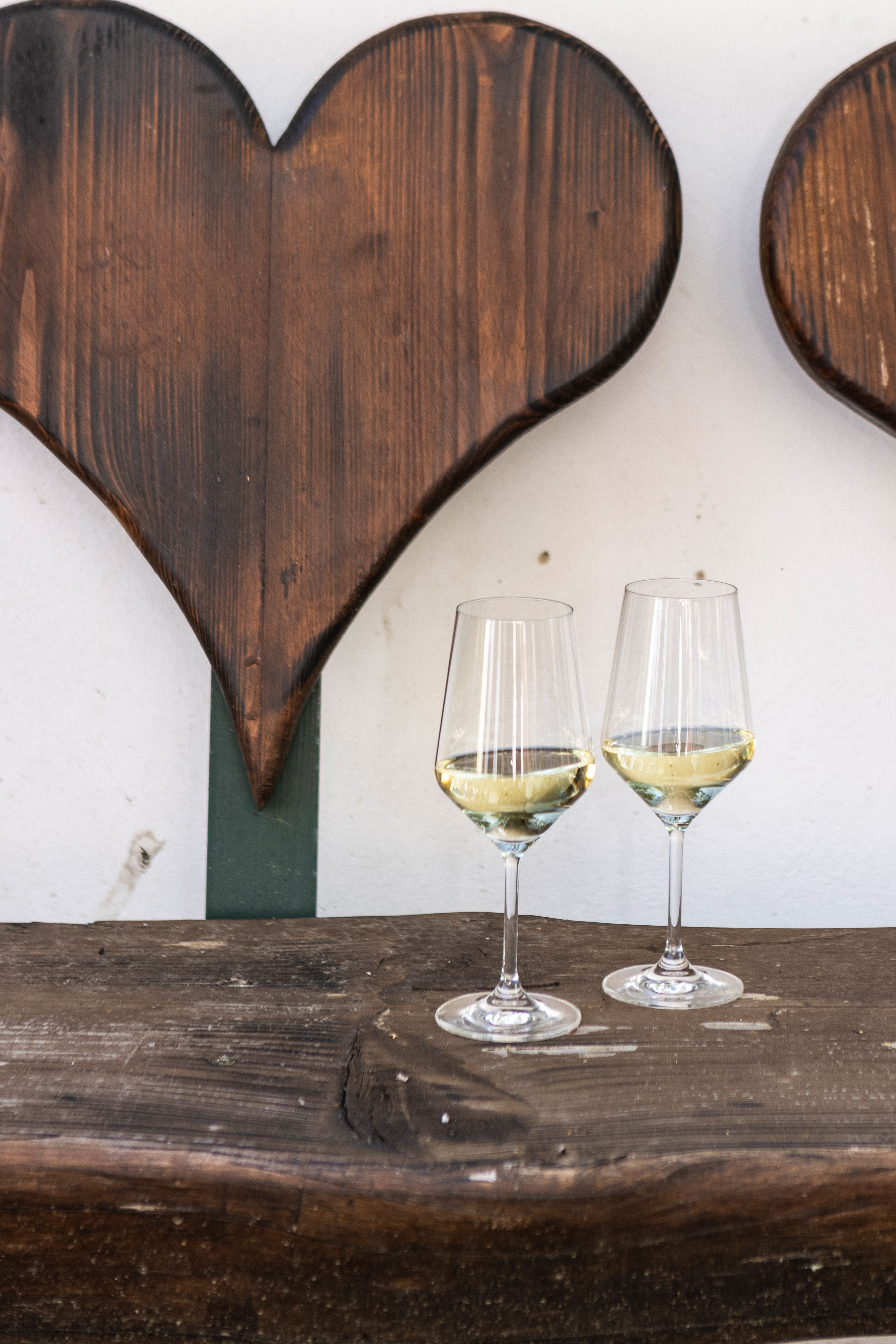 Zwei Weingläser mit Weißwein auf einem Holztisch vor einer Holzherz-Dekoration.