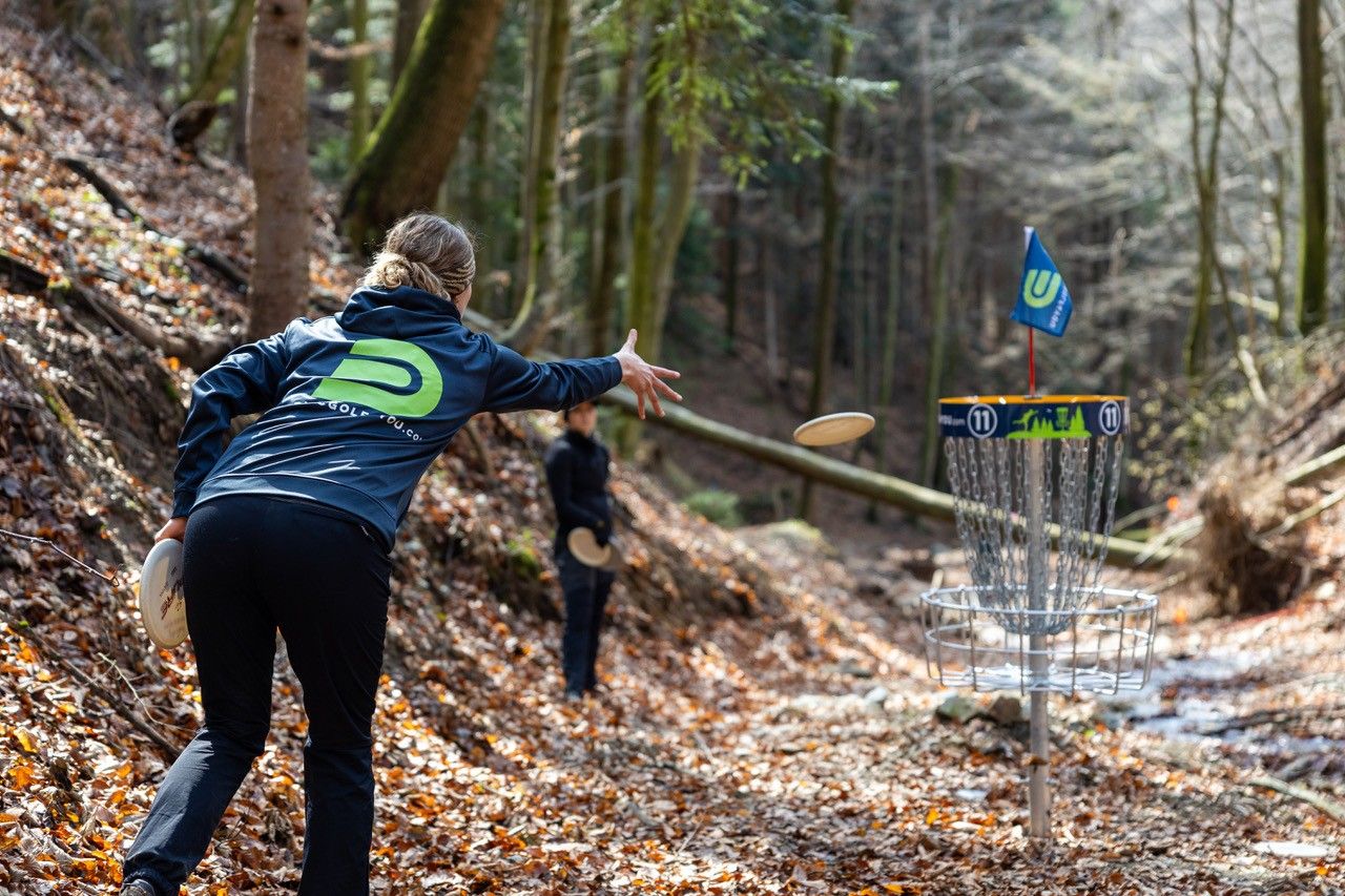 Person wirft Frisbee in Discgolf-Korb im Wald.