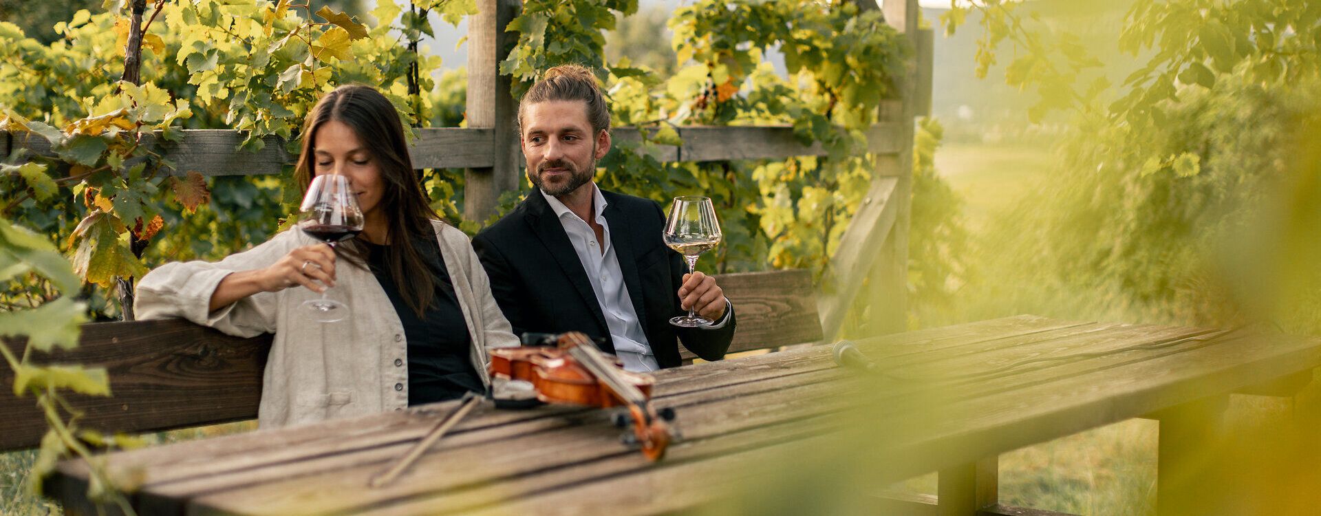 Eine Frau und ein Mann sitzen mit Weingläsern an einem Holztisch unter einer Weinlaube. Auf dem Tisch liegt eine Geige, umgeben von grünen Weinreben im warmen Abendlicht.