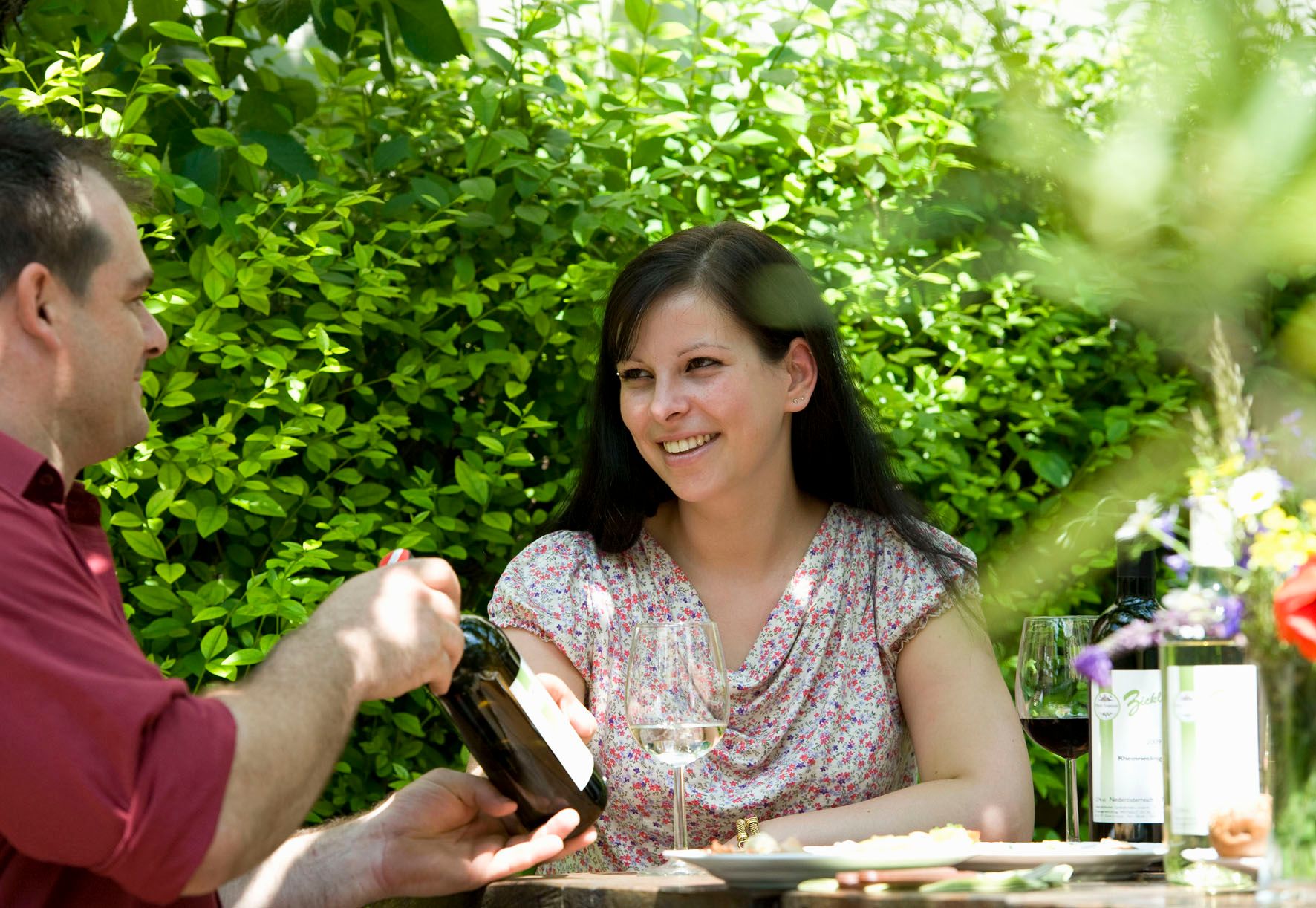 Zwei Personen sitzen im Freien an einem Tisch mit Wein und Blumen.