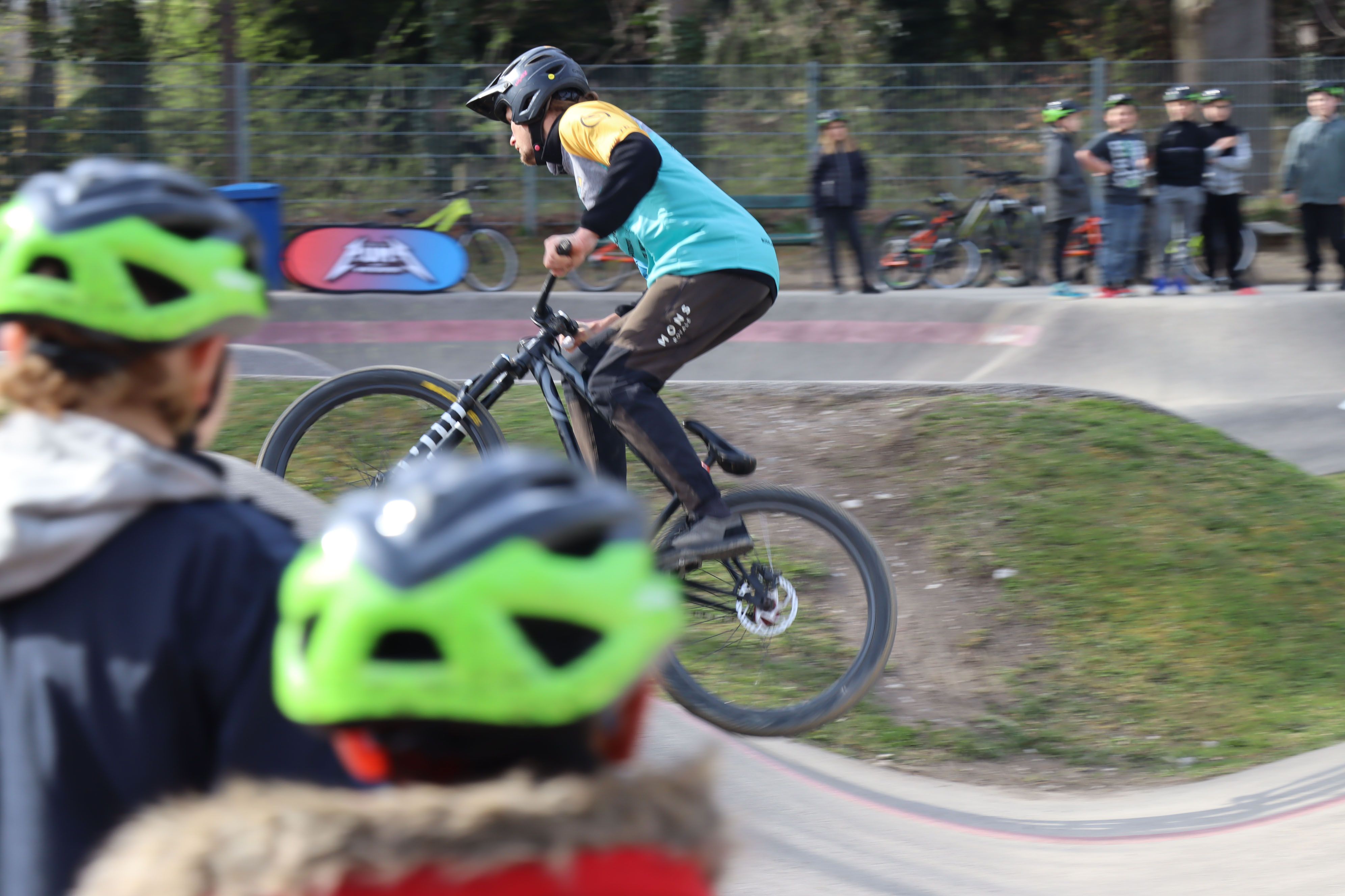 Ein Radfahrer springt auf einem Pumptrack, während Zuschauer mit grünen Helmen zuschauen.