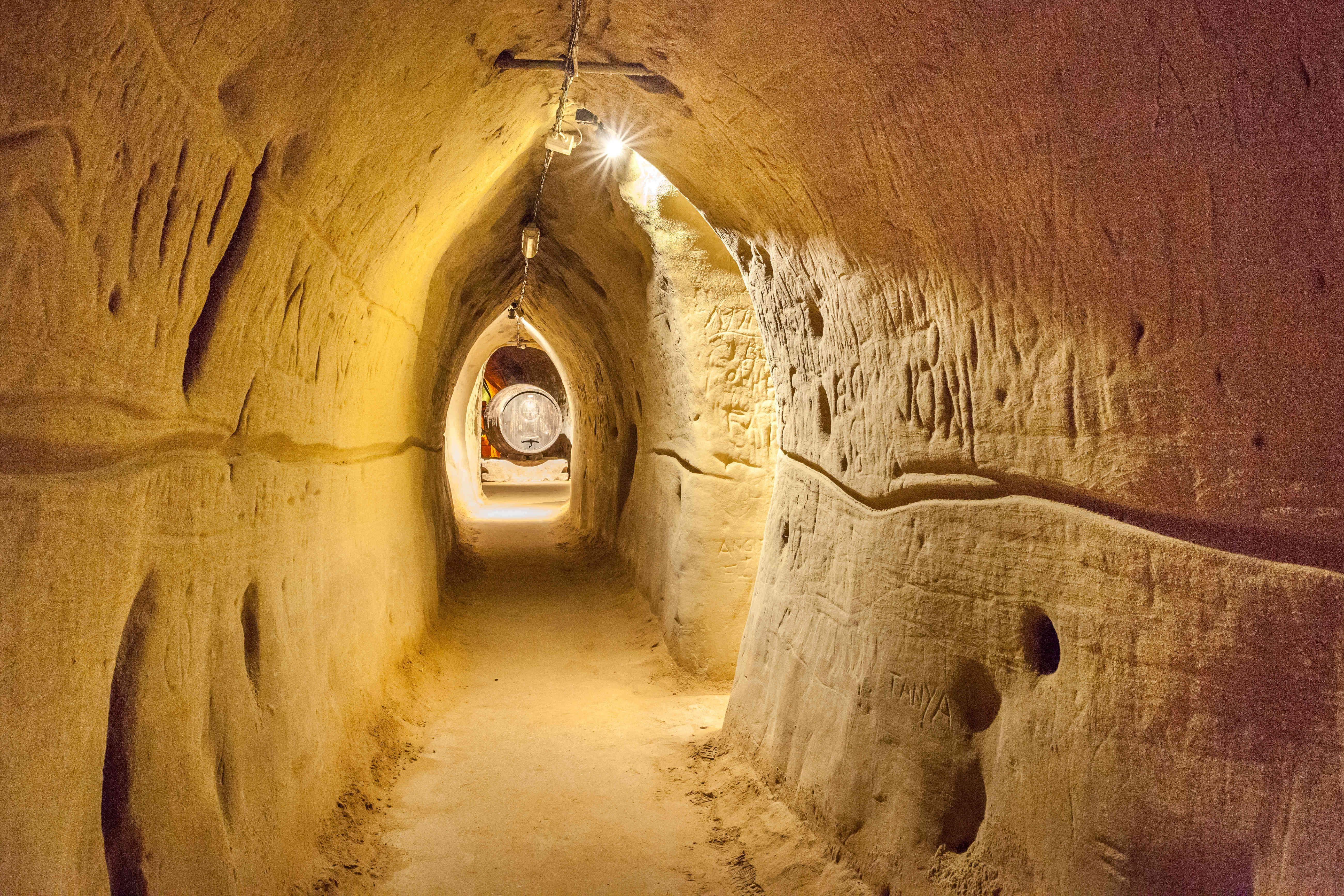 Ein beleuchteter, schmaler Tunnel mit sandfarbenen Wänden, der zu einem großen Weinfass führt.