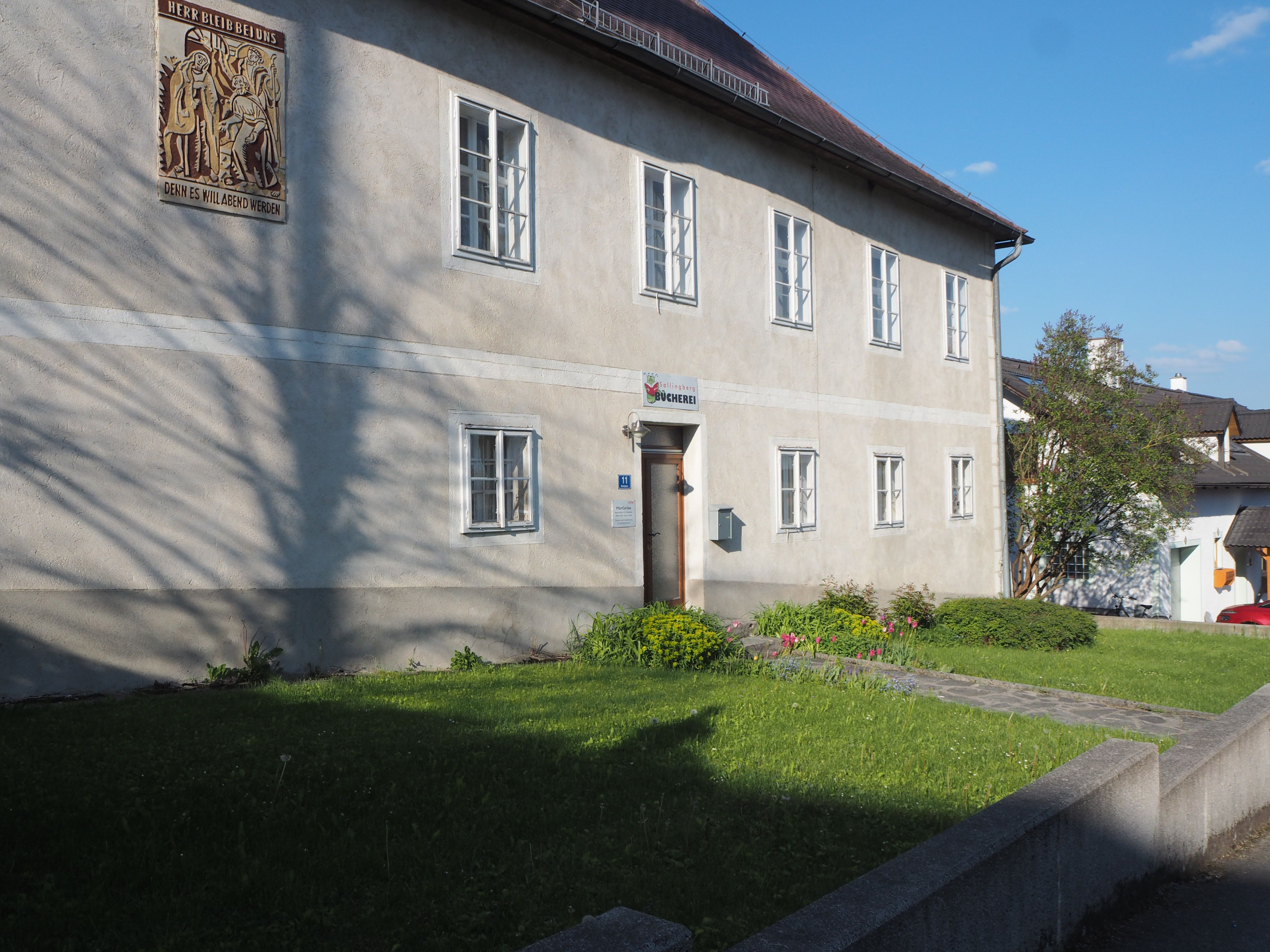 Ein zweistöckiges Gebäude mit mehreren Fenstern und einem Schild über der Tür, umgeben von einem gepflegten Garten.