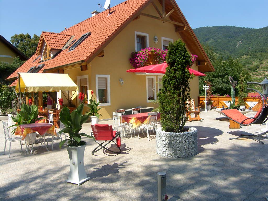 Gästehaus mit Terrasse, Tischen, Stühlen und Sonnenschirmen, umgeben von Pflanzen und Bergen im Hintergrund.