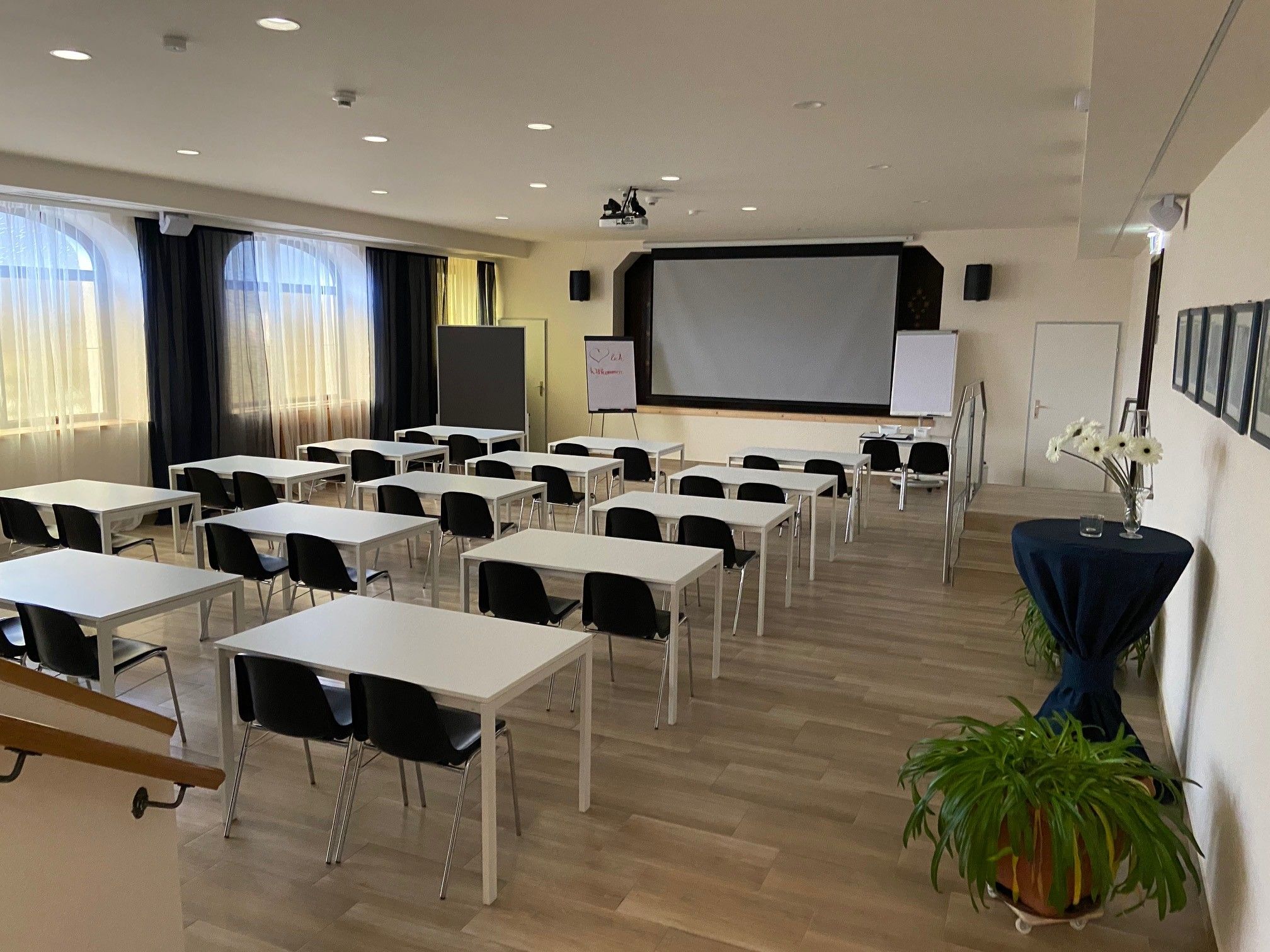 Ein leerer Seminarraum mit weißen Tischen, schwarzen Stühlen und einem großen Bildschirm vorne.