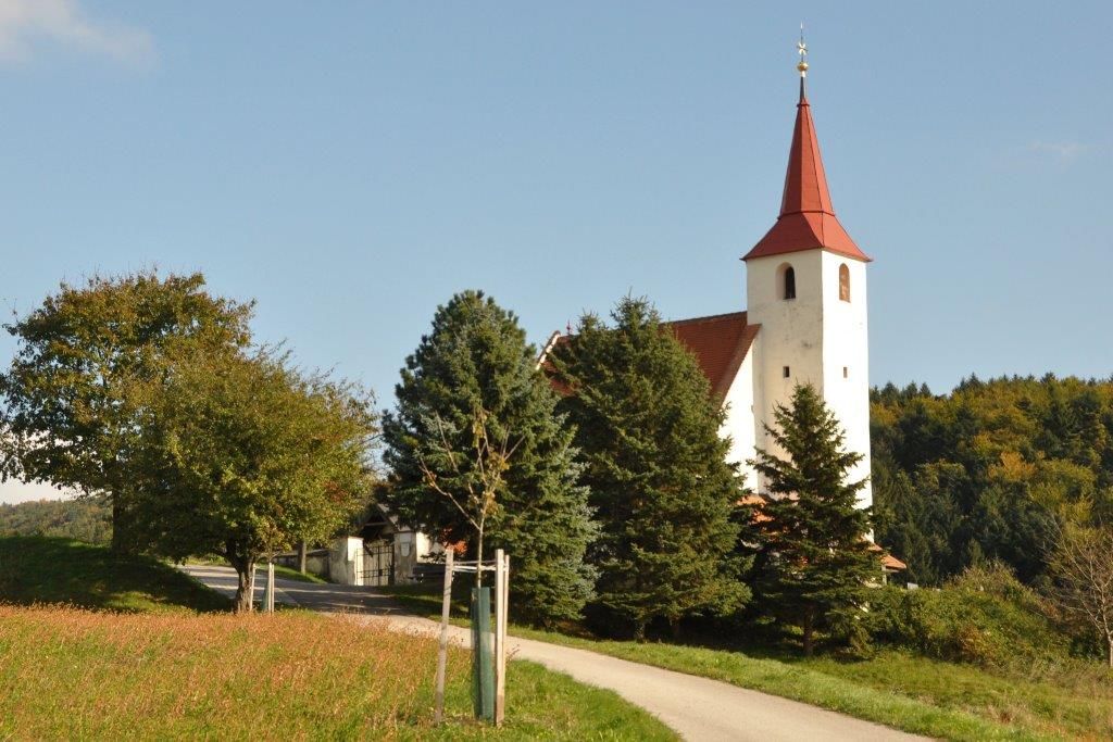 Pfarrkirche Ofenbach mit rotem Turmdach, umgeben von Bäumen und Wiesen.
