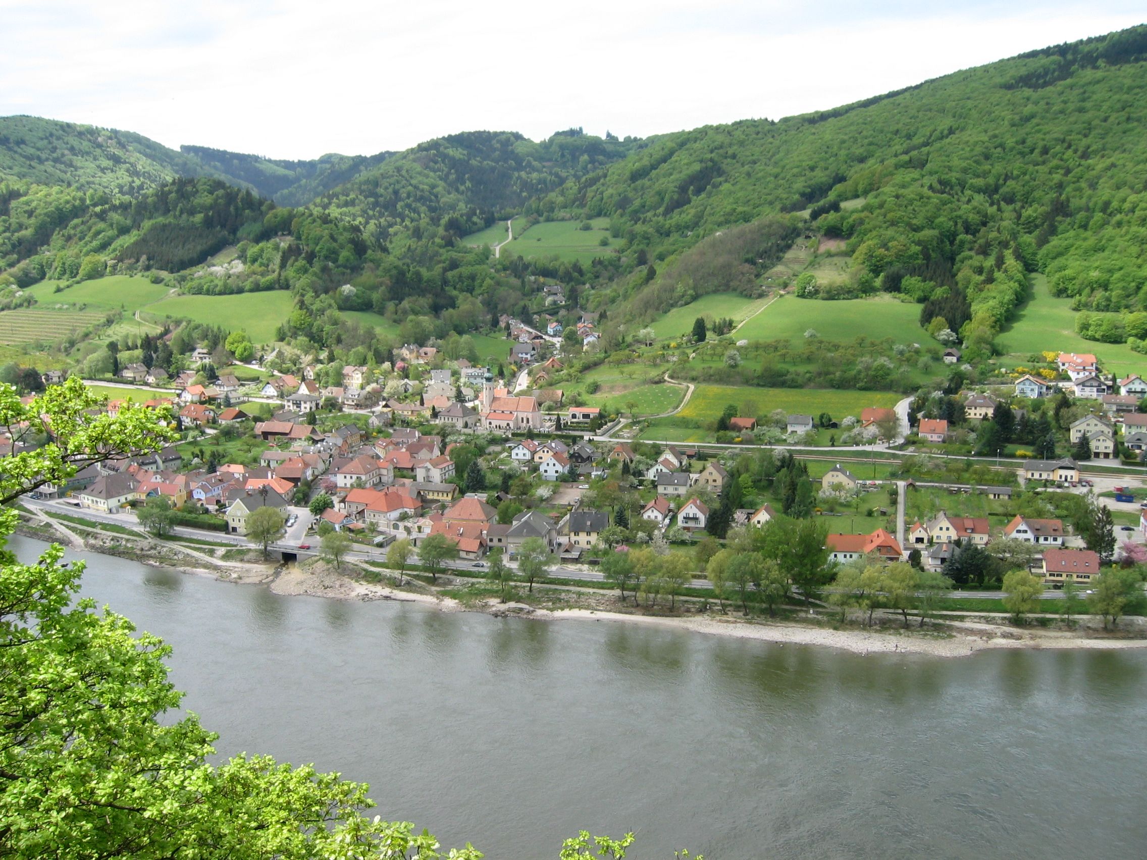 Luftaufnahme von Aggsbach mit Fluss und grünen Hügeln im Hintergrund.