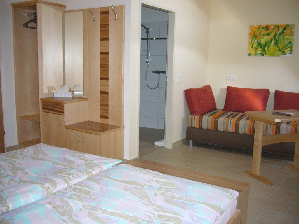 Ein gemütliches Doppelzimmer mit zwei Betten, einem Schrank, einem kleinen Tisch und einer Couch. Im Hintergrund ist ein Badezimmer zu sehen.