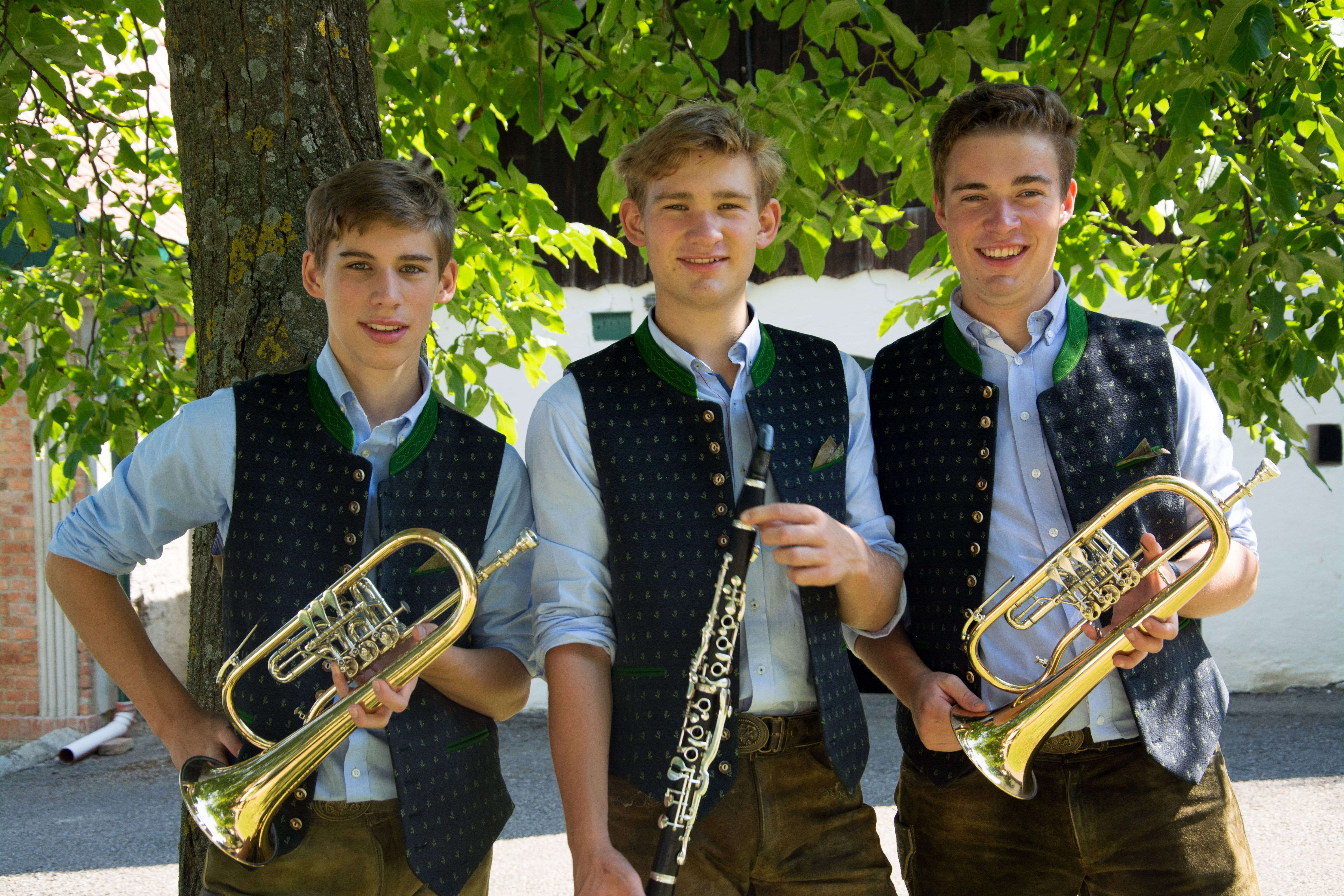 Drei junge Männer in traditioneller Kleidung mit Blasinstrumenten vor einem Baum.