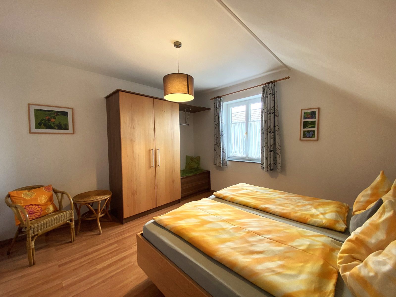 Schlafzimmer mit Doppelbett, Holzschrank, Korbstuhl und Fenster mit Vorhängen.