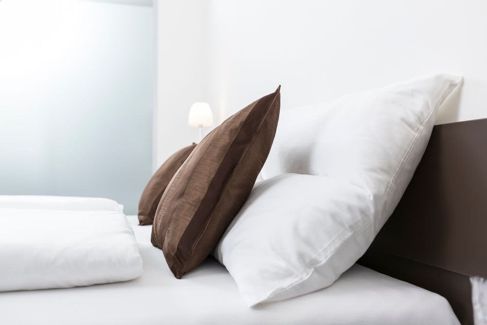 Ein Bett mit weißen Kissen und braunen Zierkissen, im Hintergrund eine Lampe.