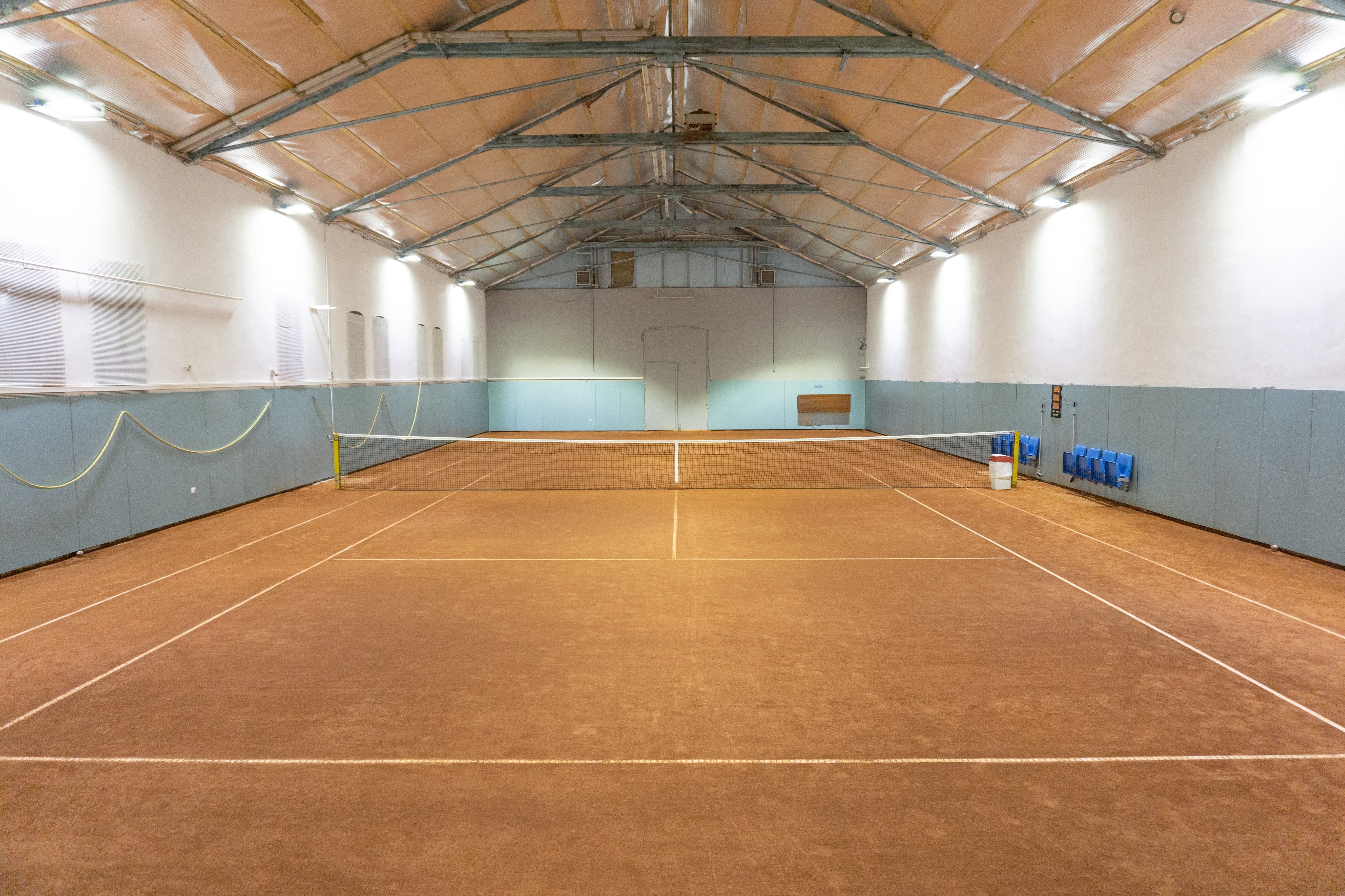Innenansicht eines leeren Tennisplatzes in einer Halle mit braunem Bodenbelag und blauen Wänden.