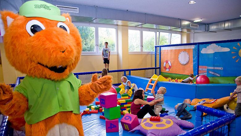 Kinder spielen in einem bunten Indoor-Spielbereich mit einem großen, orangefarbenen Maskottchen.