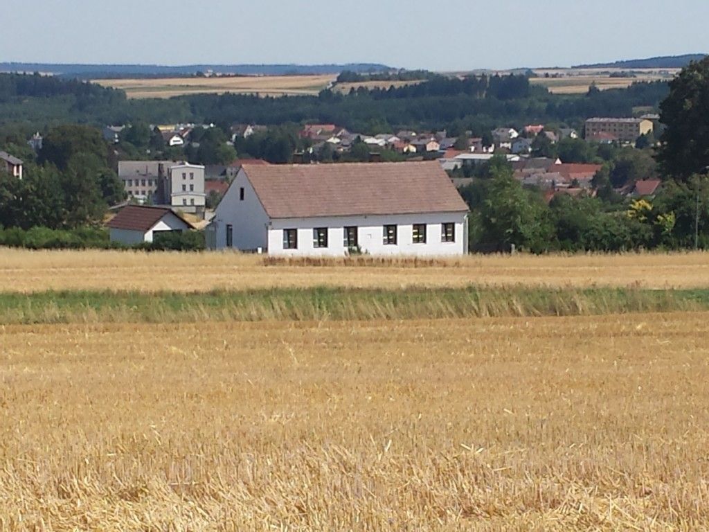 Ein weißes Haus mit rotem Dach steht in einem Feld, im Hintergrund eine Stadt und Wälder.