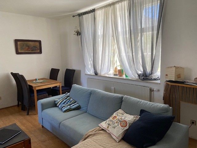 Ein Wohnzimmer mit einem blauen Sofa, einem Holztisch mit Stühlen und einem Fenster mit weißen Vorhängen.