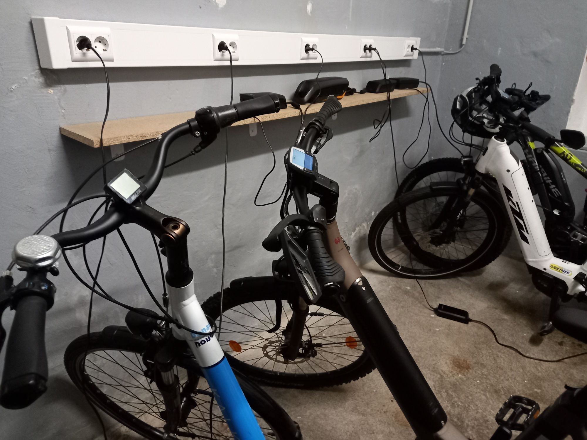 Drei E-Bikes in einer Garage, die an einer Steckdosenleiste aufgeladen werden.