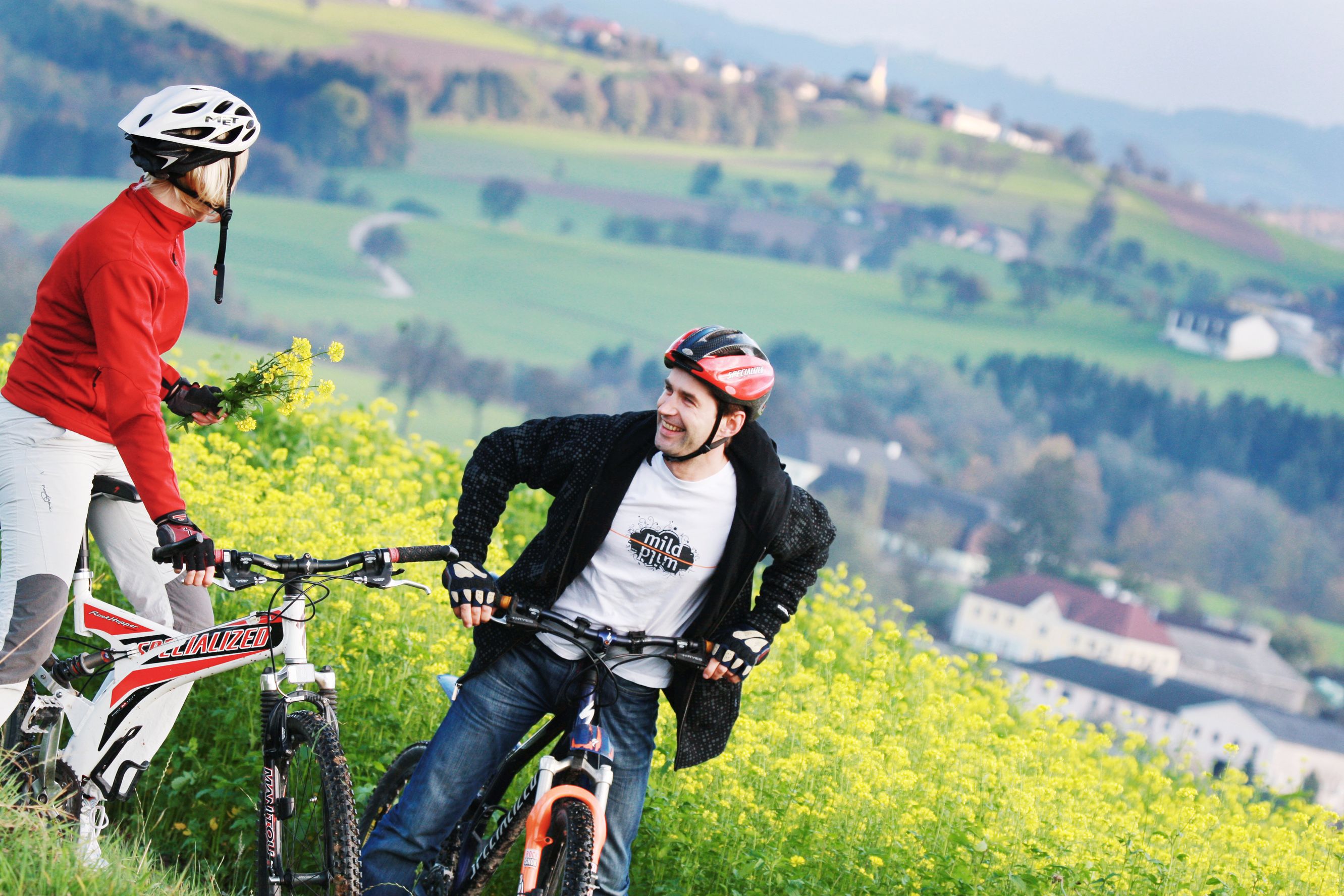 Zwei Radfahrer mit Helmen stehen auf einem blühenden Feld mit grüner Landschaft im Hintergrund.