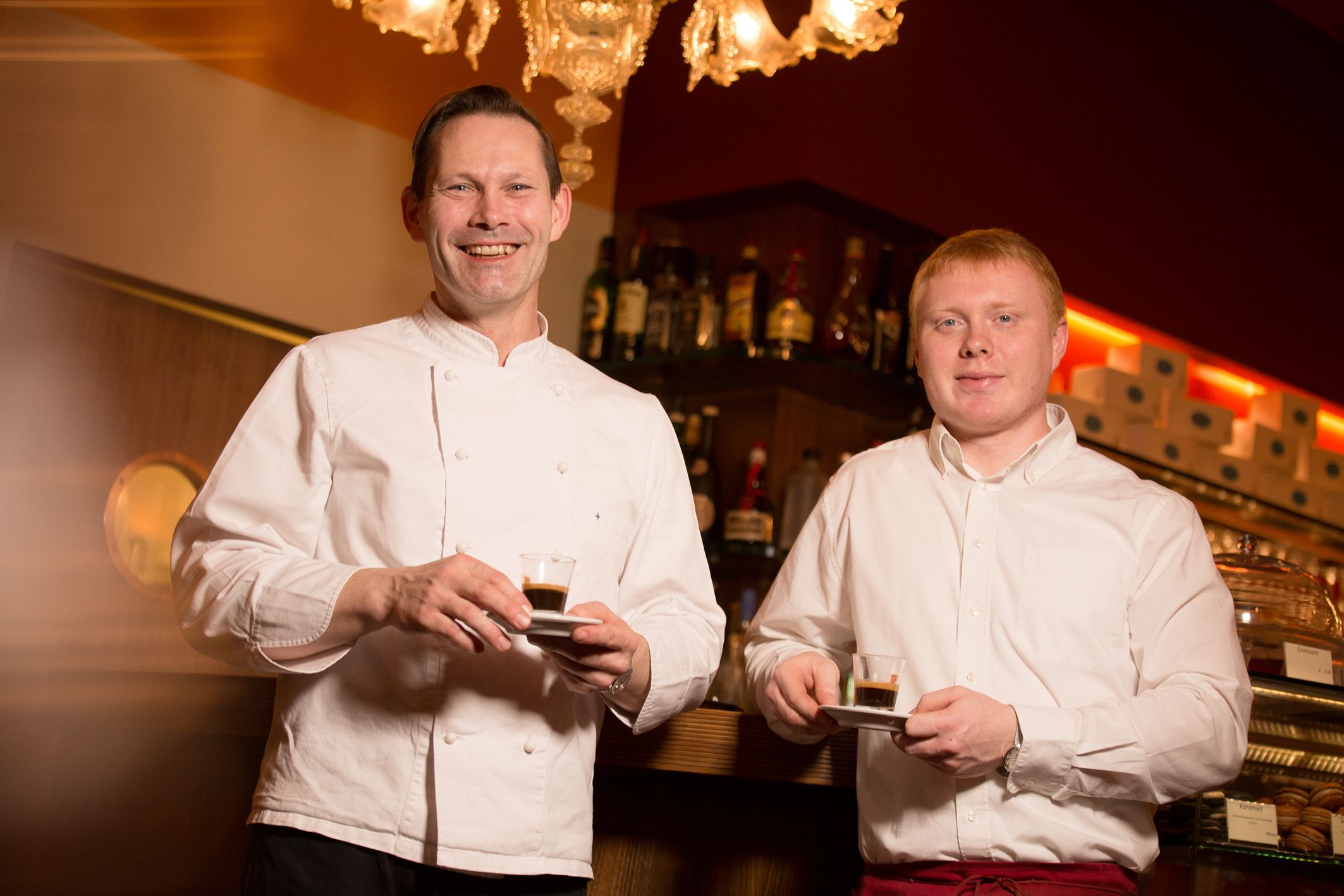 Zwei Männer in einem Café halten Espressotassen und lächeln in die Kamera.