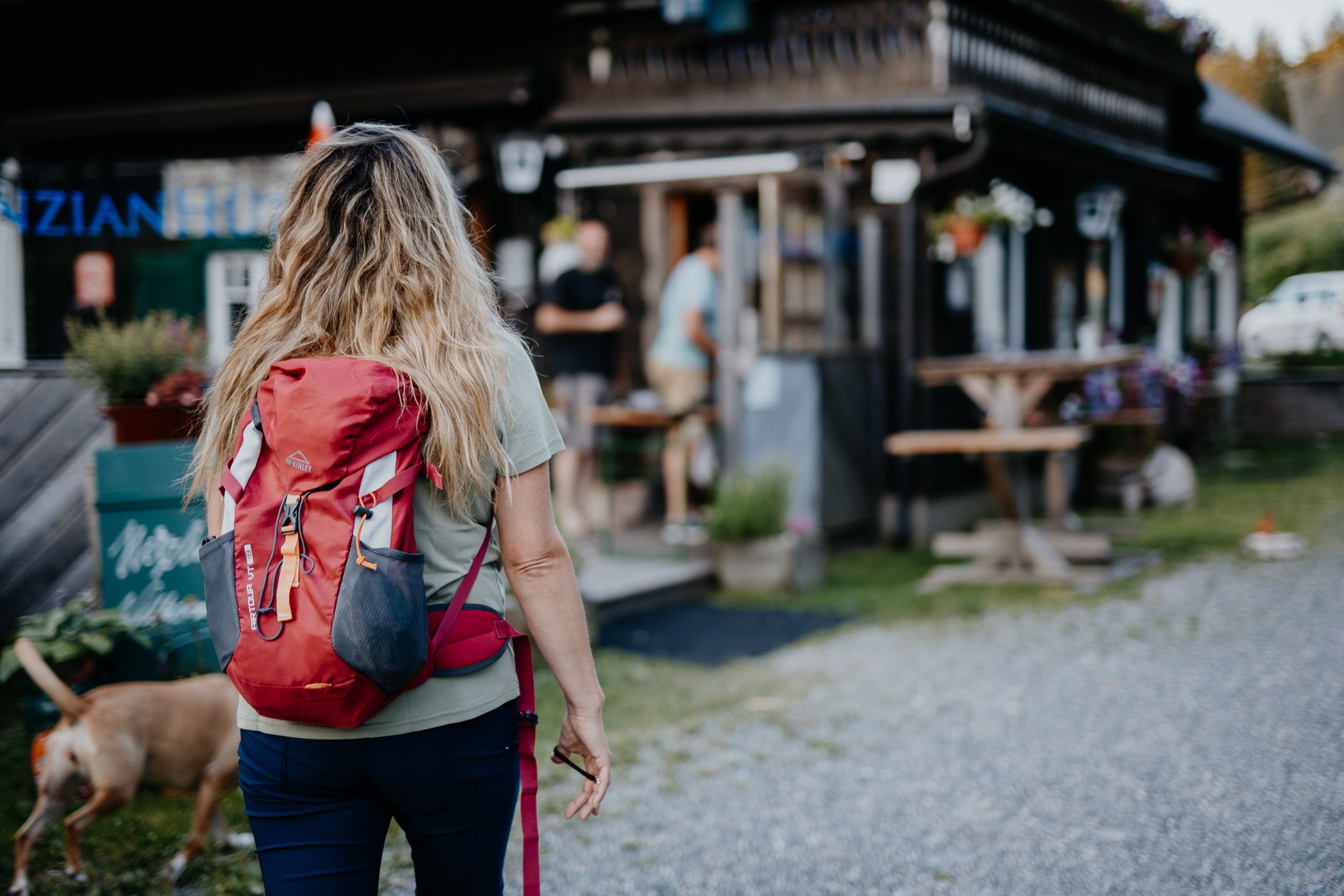 Frau mit rotem Rucksack und Hunde geht auf eine Berghütte zu.
